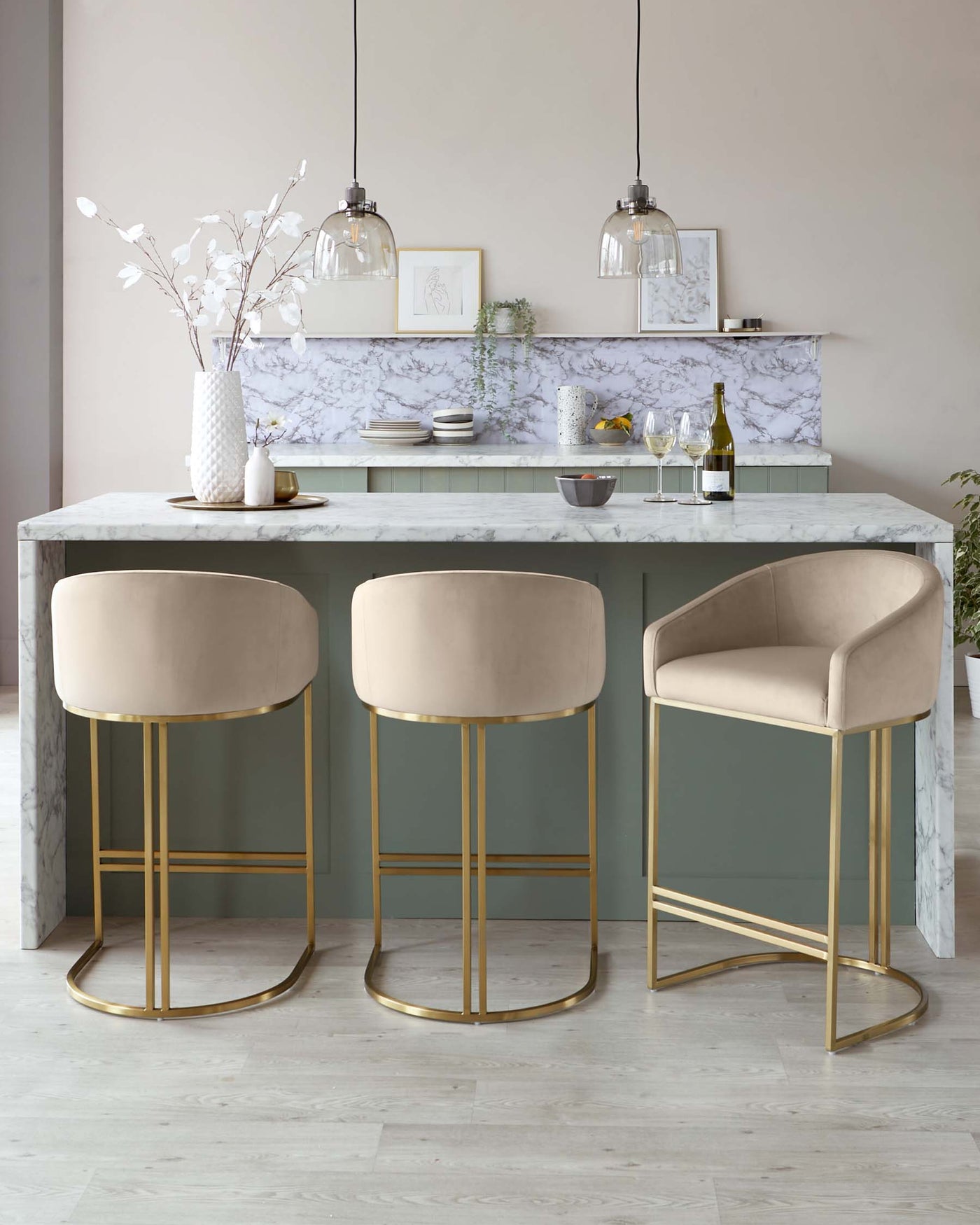 Elegant modern bar stools with blush pink upholstery and sleek gold metal frames, showcasing a circular seat design and a semi-open backrest, placed against a kitchen island with a marble countertop.