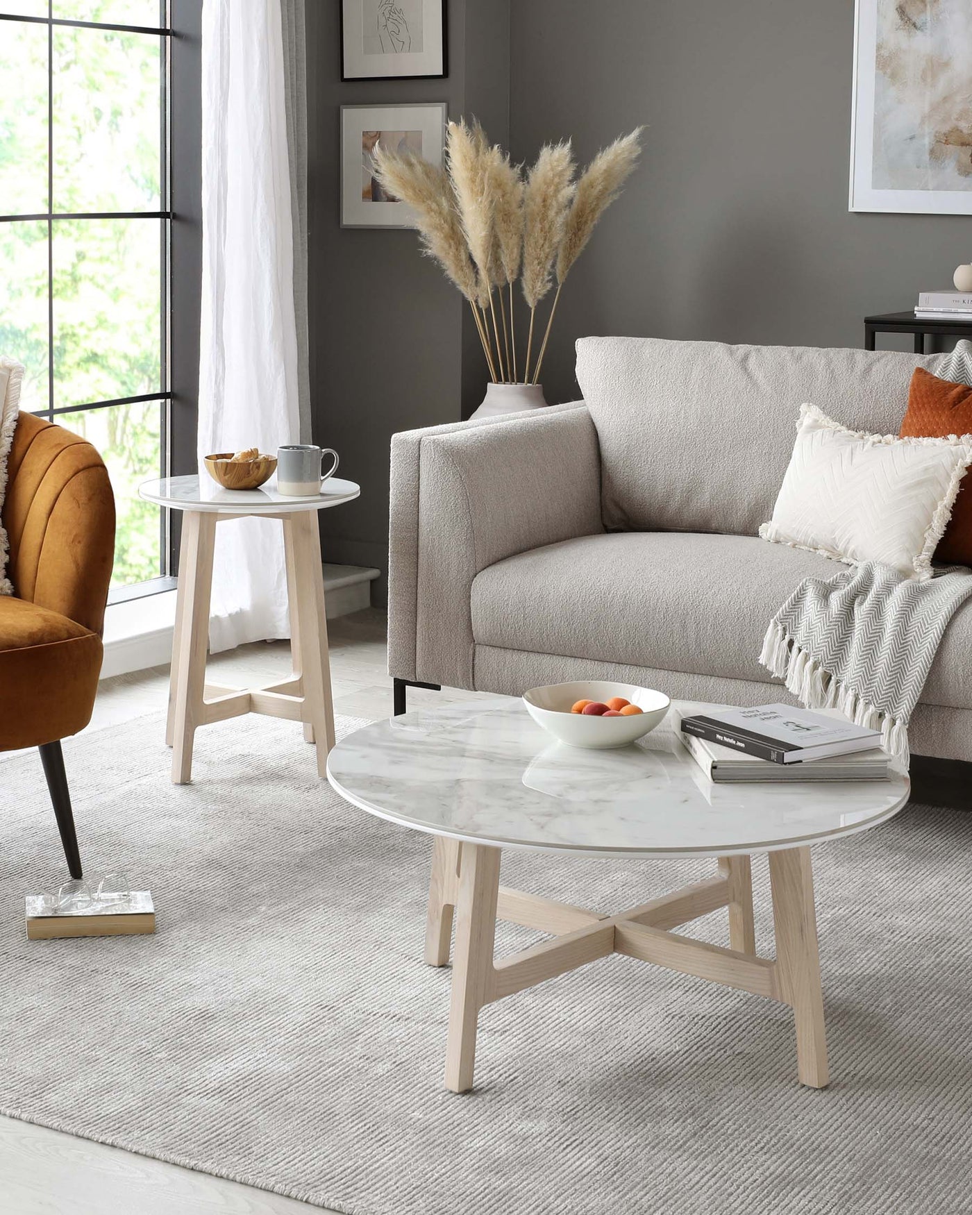 Modern living room setup featuring a light grey fabric sofa with plush cushions, a mid-century modern amber velvet armchair with a curved back and angled wooden legs, and a round white marble coffee table with a unique wooden cross-base design. A simplistic round white side table with three wooden legs is also present, completing the contemporary aesthetic. The space is tied together with a textured grey area rug.
