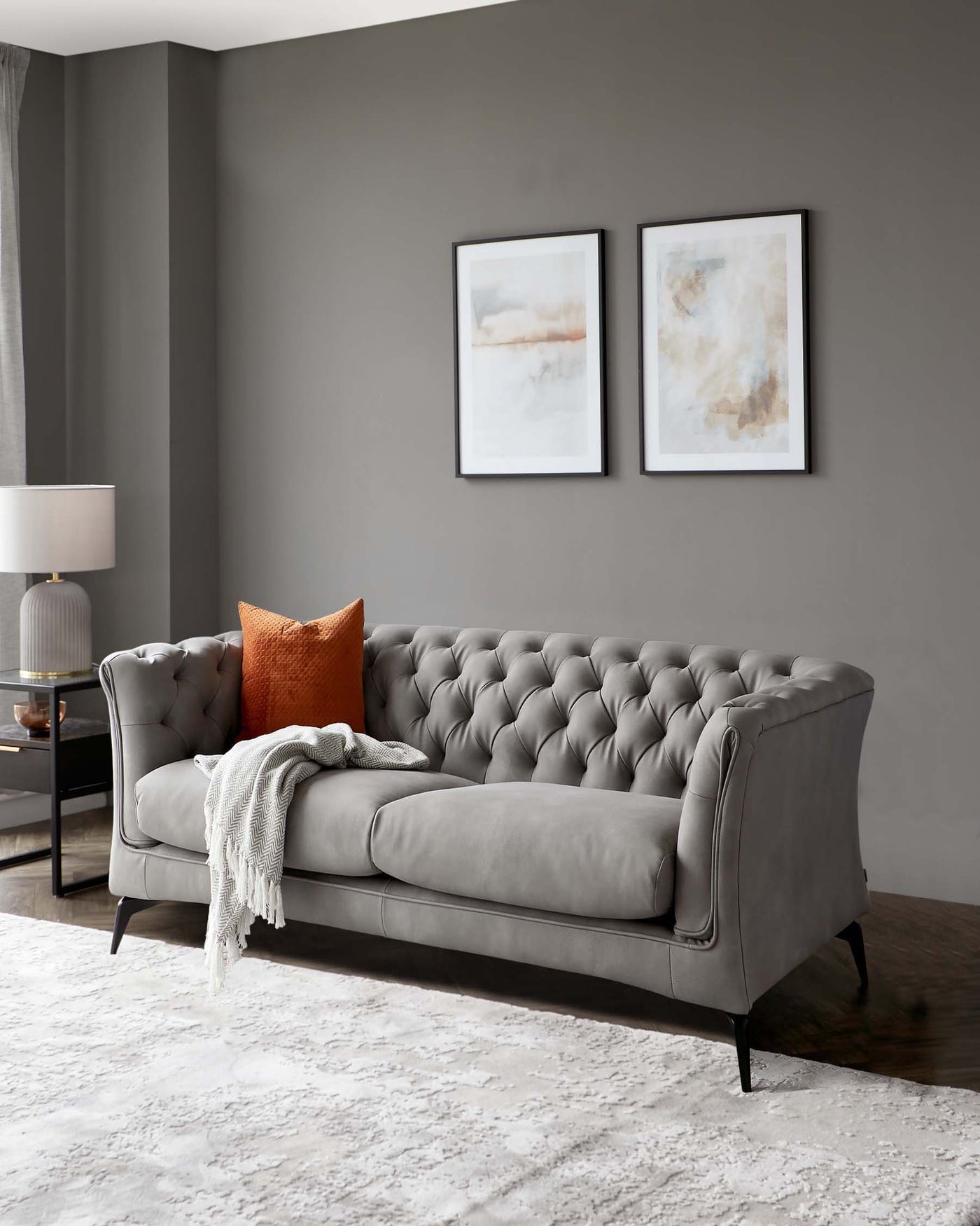 Elegant grey tufted sofa with rolled arms and black wooden legs, adorned with a burnt orange velvet cushion and a light grey woven throw blanket.