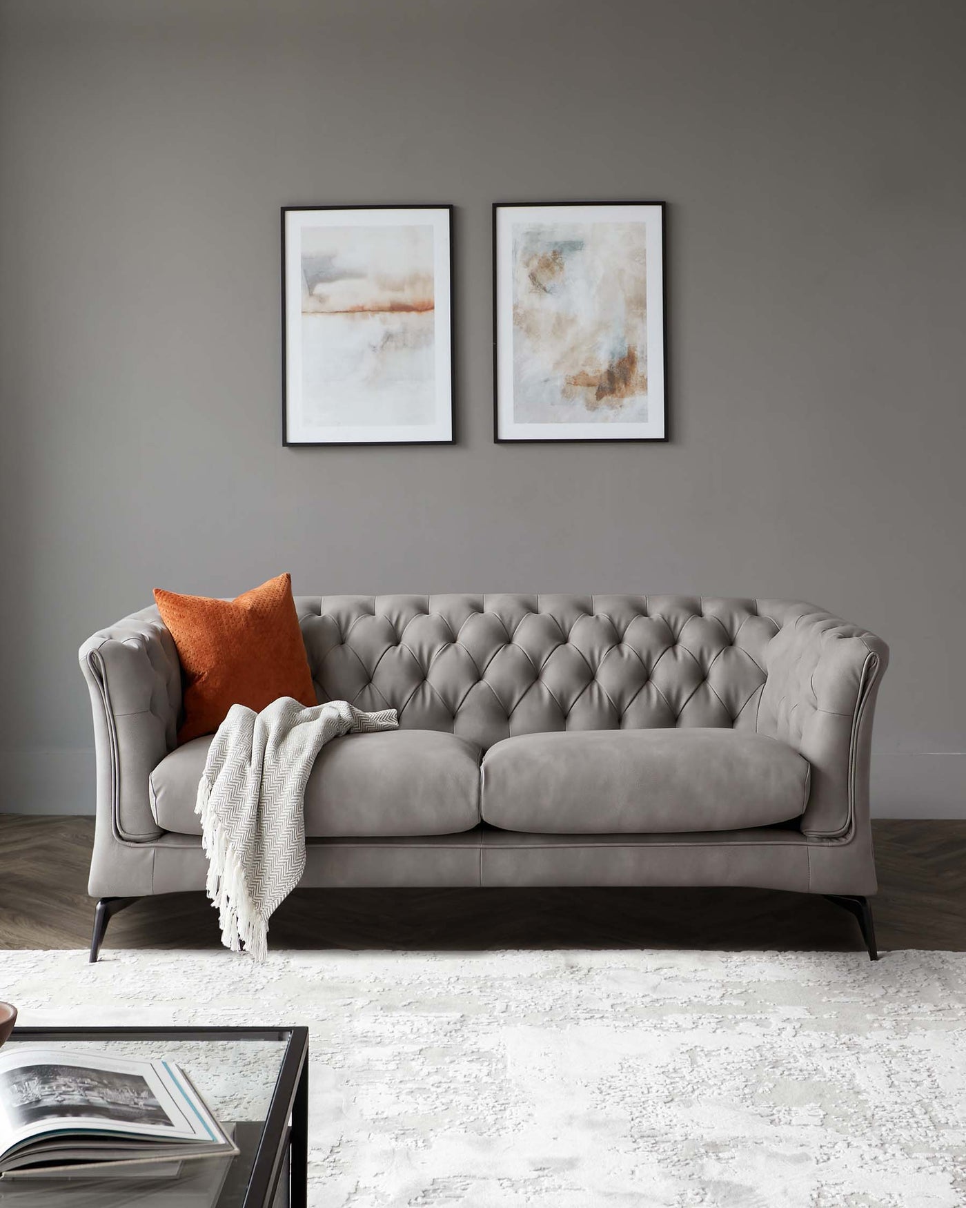 Elegant grey tufted sofa with rolled arms and dark wooden legs, accented with an orange decorative pillow and a white throw blanket.