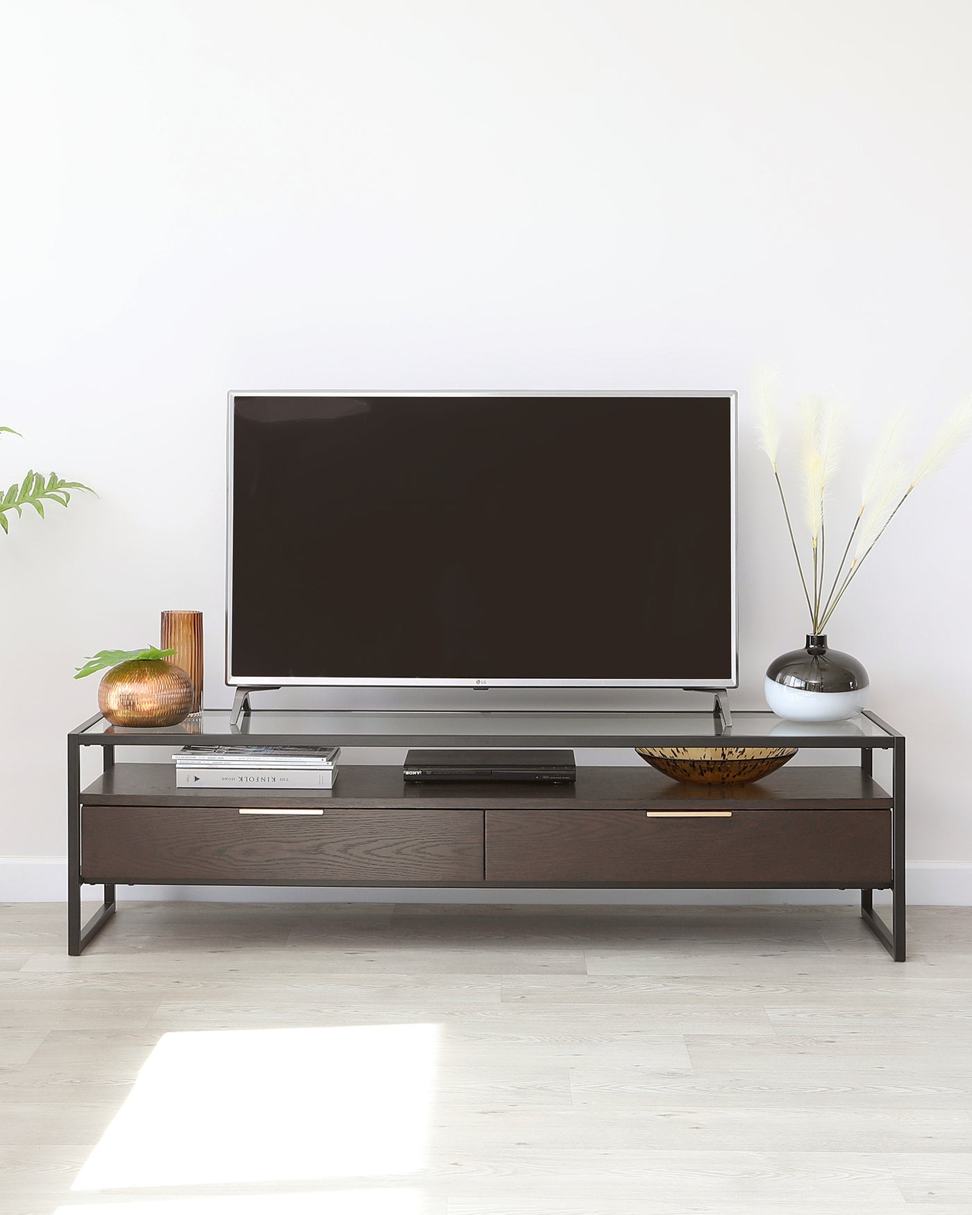 Modern minimalist TV stand with a dark wood finish and metal frame, featuring two drawers and an open shelf for storage, displayed in a bright room with decorative items.