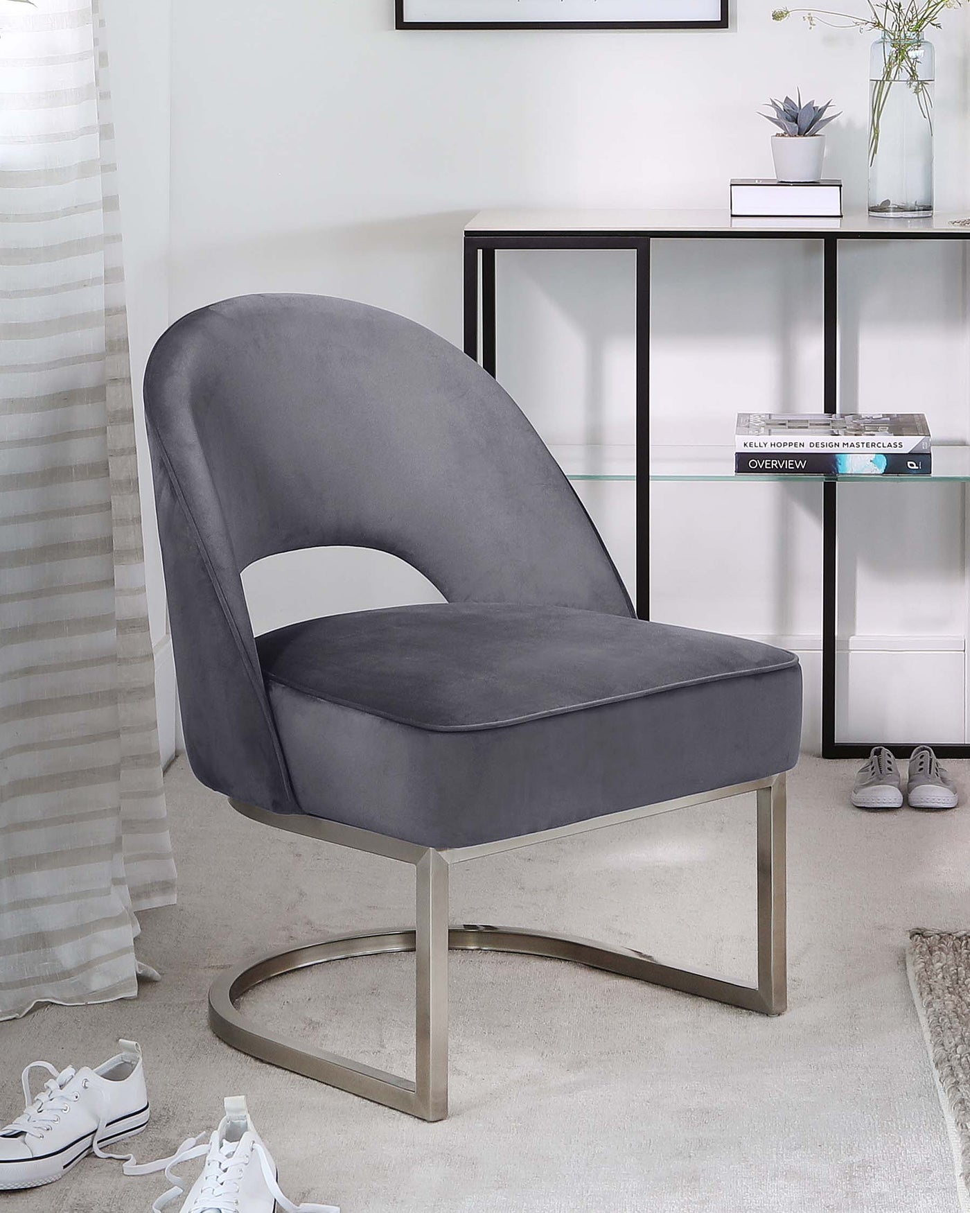 Modern grey velvet accent chair with curved backrest and unique silver metal base, positioned beside a black-framed shelving unit with glass shelves displaying books and decorative items, set against a neutral-toned room with white walls and light carpet flooring.