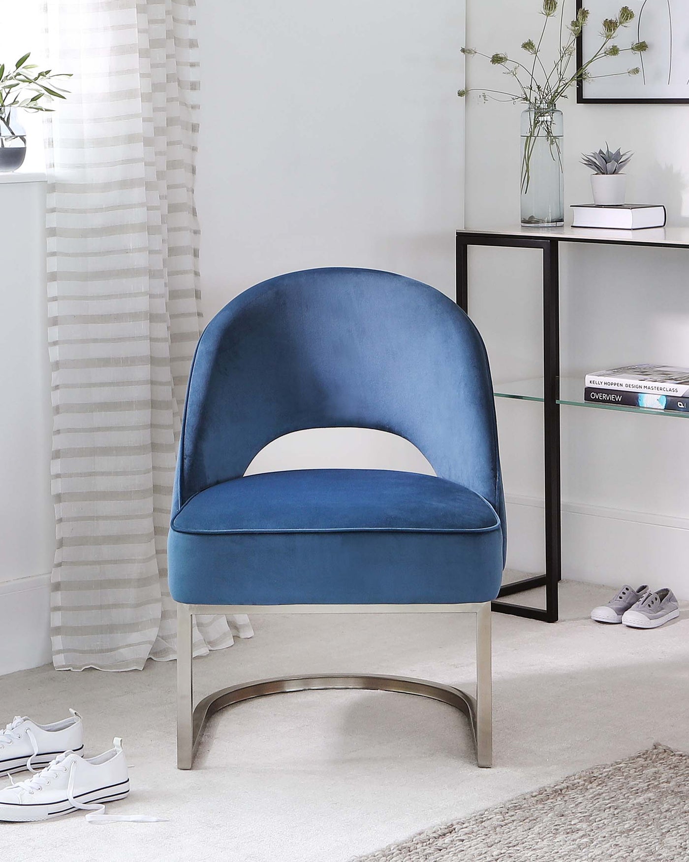 Modern blue velvet accent chair with a curved high-back design and a metal silver base. Adjacent to the chair is a contemporary glass and metal frame side table with two shelves housing books and decorative items.