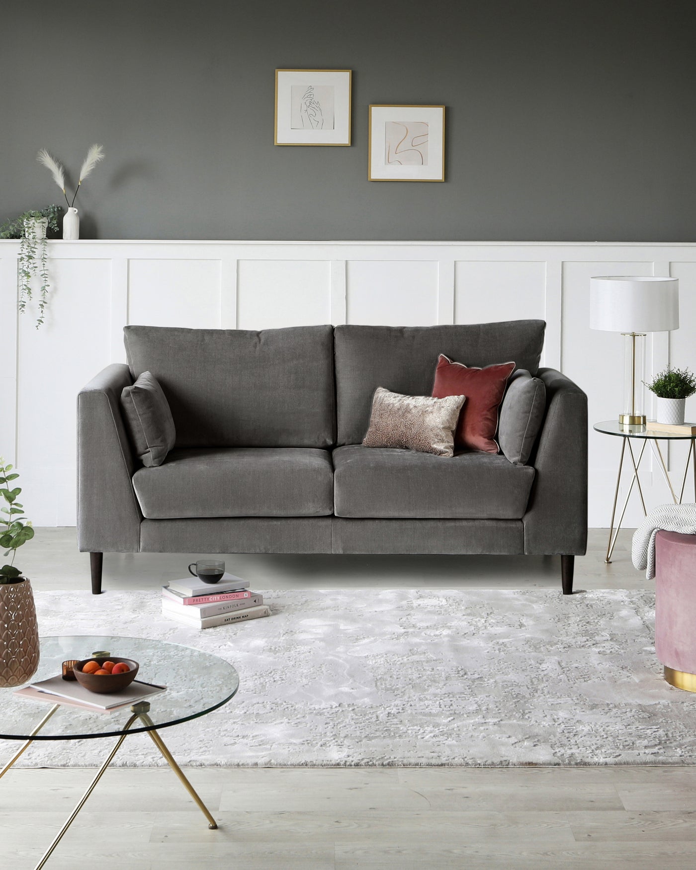 Elegant living room furniture arrangement featuring a modern, grey fabric sofa with plush cushioning and clean lines, accompanied by a gold-framed circular glass coffee table with a unique crossed-leg design. A chic side table with a white round tabletop and golden legs is set beside a contemporary white floor lamp with a cylindrical shade. A textured light-coloured area rug anchors the space, adding a cosy layer to the wooden flooring.