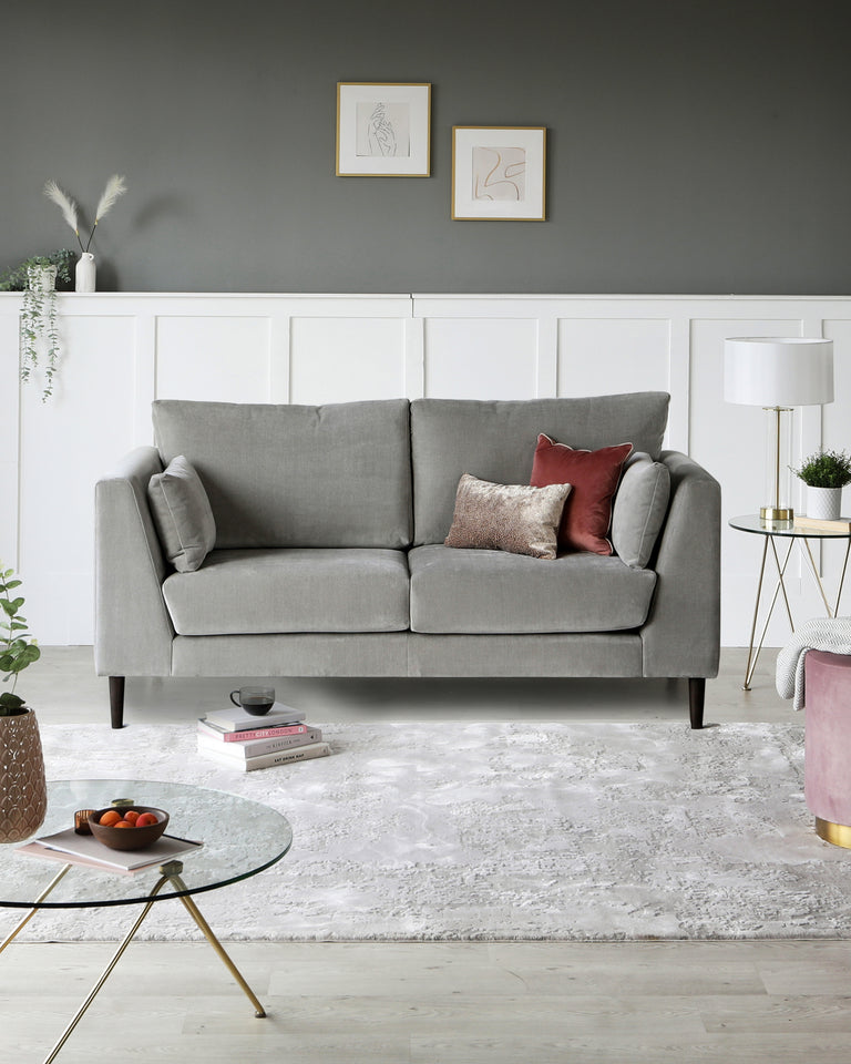 A contemporary living room setup featuring a grey upholstered two-cushion sofa with a tufted backrest and sleek armrests. A round glass coffee table with golden metal legs sits on a textured white area rug, accompanied by a white lamp with a gold base on a small round side table with a marble top and golden legs. A pink cylindrical ottoman with a gold base adds a pop of colour to the scene.