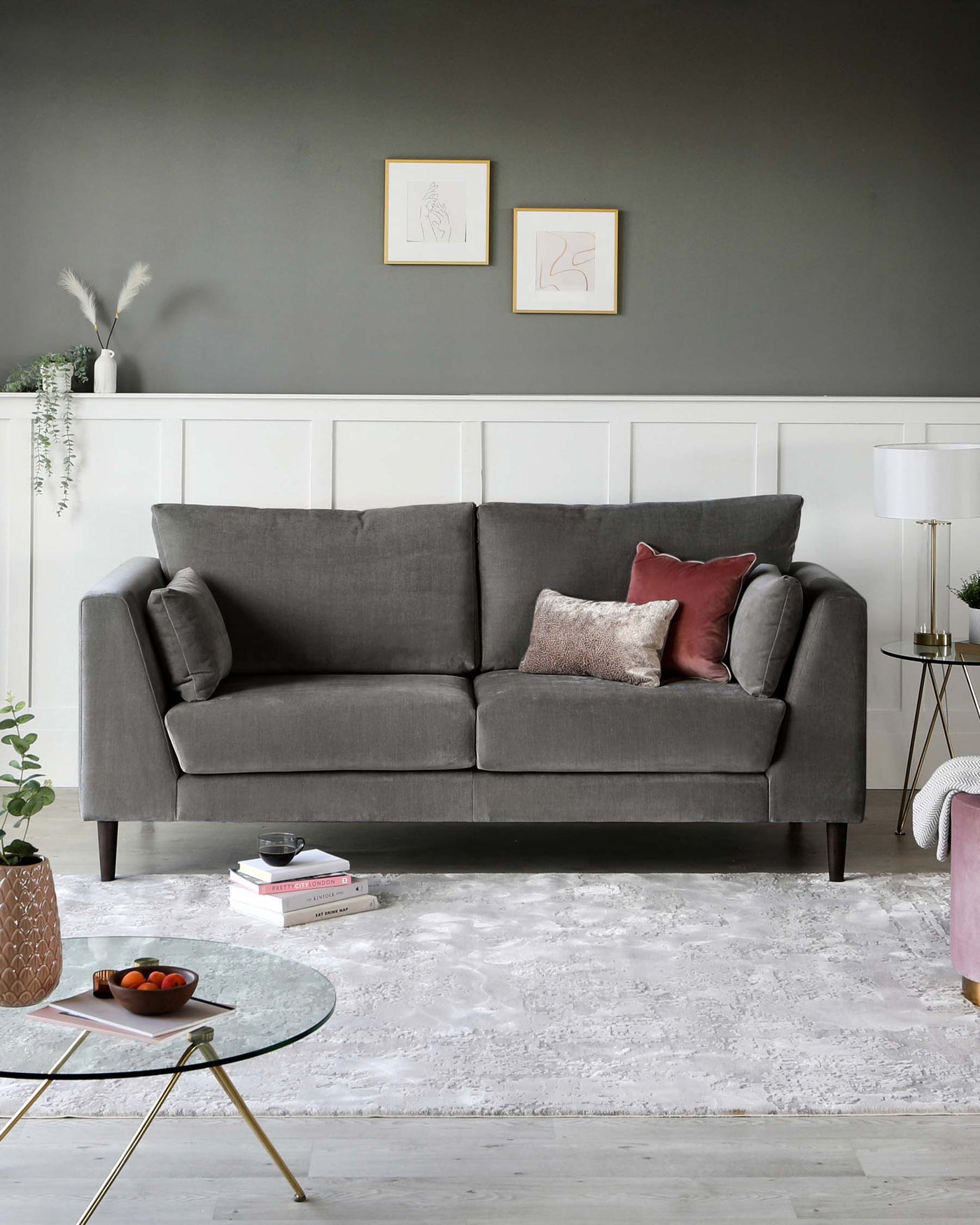 Elegant living room setup featuring a contemporary grey upholstered three-seater sofa with cushioned armrests, two grey throw pillows, and one each of dusty rose and metallic textured decorative cushions. A round glass-top side table with a gold-finished geometric base rests on a plush white area rug, accompanied by a small planter and books for accent. A modern white lamp with a clear stand and white shade complements the side table.