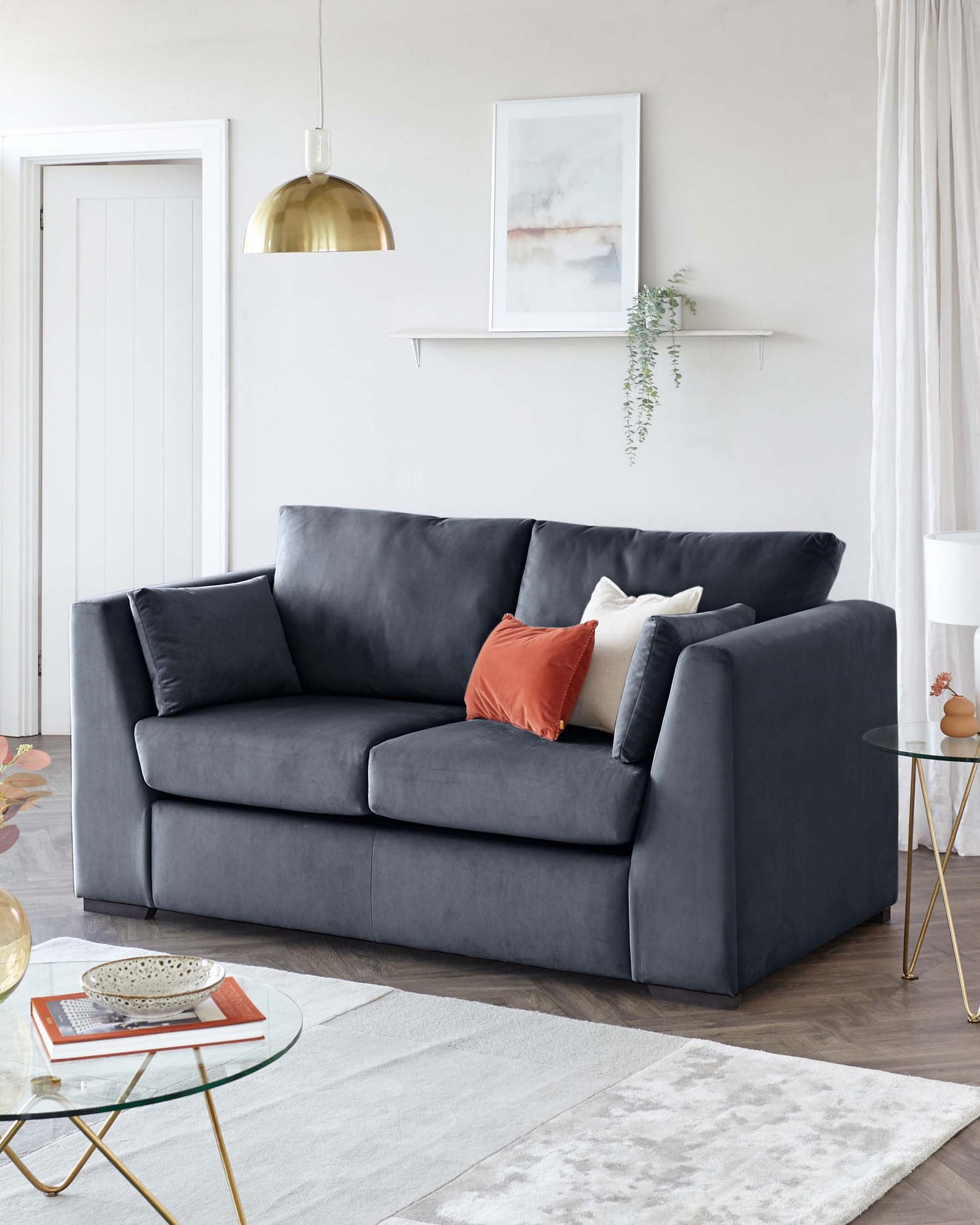 A contemporary charcoal grey upholstered sofa with plush cushions and a clean-lined silhouette. In front of the sofa is a modern round glass-top coffee table with a gold geometric frame. The room is accented with a white and grey patterned area rug.