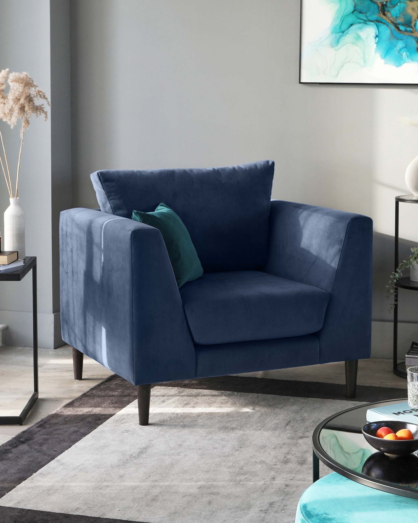 A contemporary navy blue velvet armchair with a clean, square design, featuring plush cushions and dark wooden legs. The chair is accented with a teal throw pillow and is positioned on a two-tone grey area rug. In the background, there's a small side table with a vase and dried pampas grass, adjacent to a glass-topped coffee table with a bowl of fruit. The room is well-lit, and there's a large abstract painting with blue and green hues on the wall.