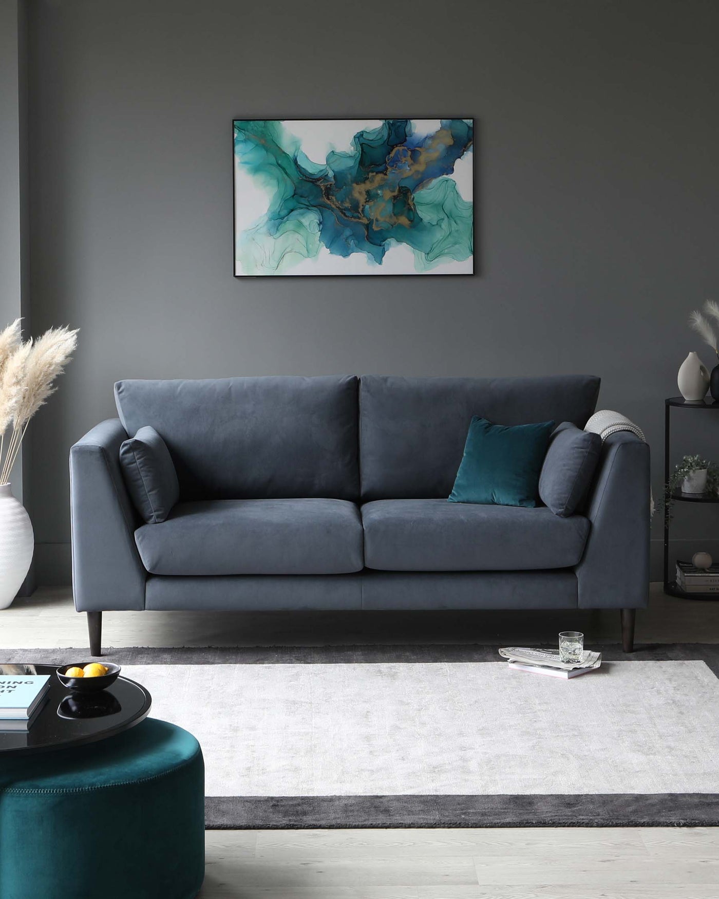 A modern living room with a three-seater plush velvet sofa in a shade of dark grey, featuring deep cushioned seats and structured armrests. In the foreground is a round, teal-coloured velvet ottoman and a rectangular rug with a faded pattern in white and grey tones. A sleek dark round coffee table with a reflective surface sits atop the rug. The room is decorated in a contemporary style with a muted colour palette.