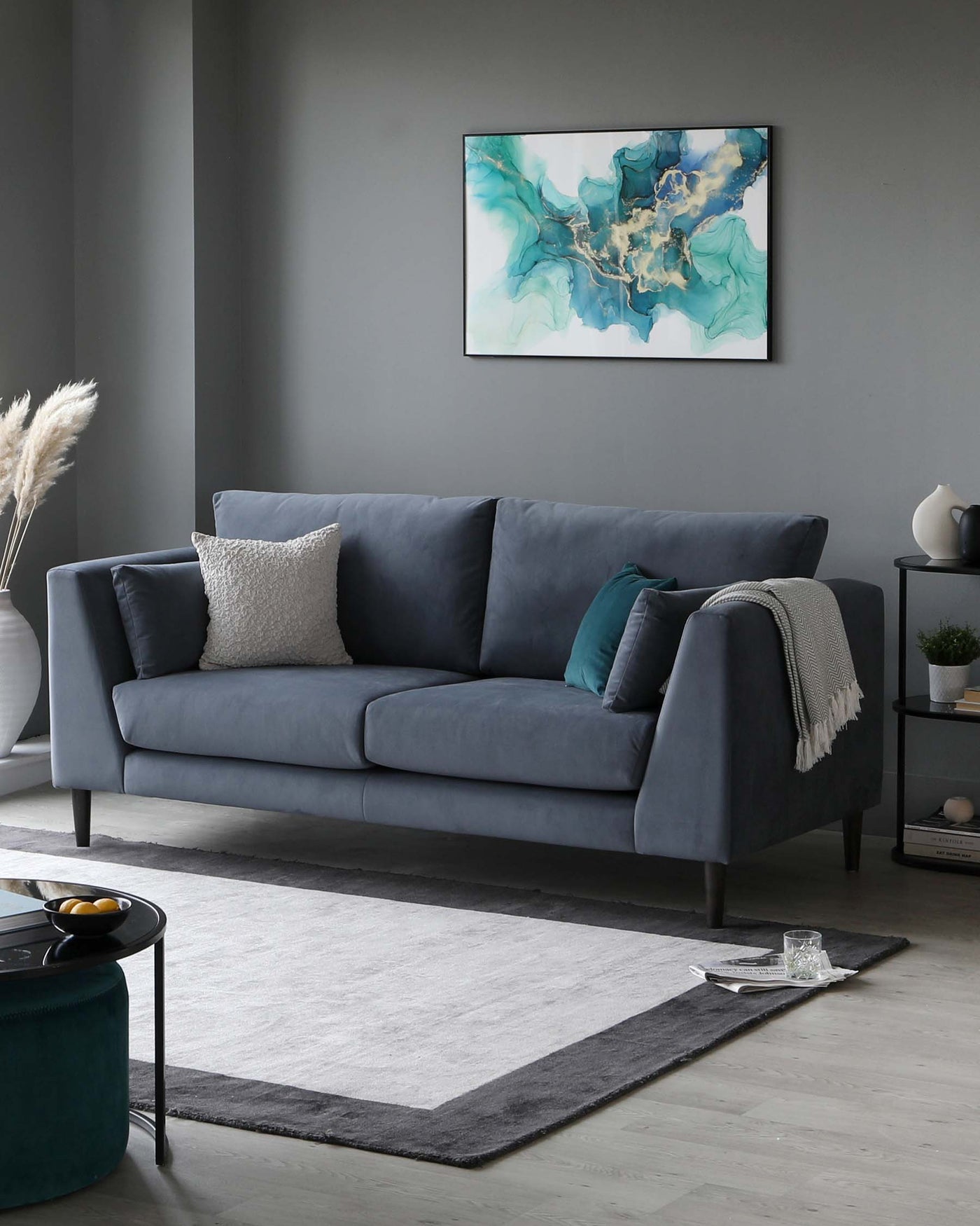 Modern three-seater sofa with plush cushions in a muted grey upholstery, accented with a mix of throw pillows in white and teal. A minimalist black metal round coffee table sits adjacent to the sofa, complemented by a dark teal round ottoman with a velvet-like finish. An off-white and grey area rug anchors the space, and a sleek black side table with shelves holds small decorative items.