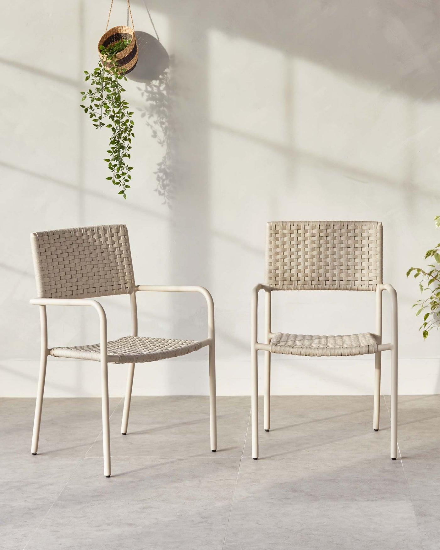 Two modern armchairs with white metal frames and woven taupe seats and backrests, showcased on a grey tiled floor against a white wall with shadow patterns from hanging plants.