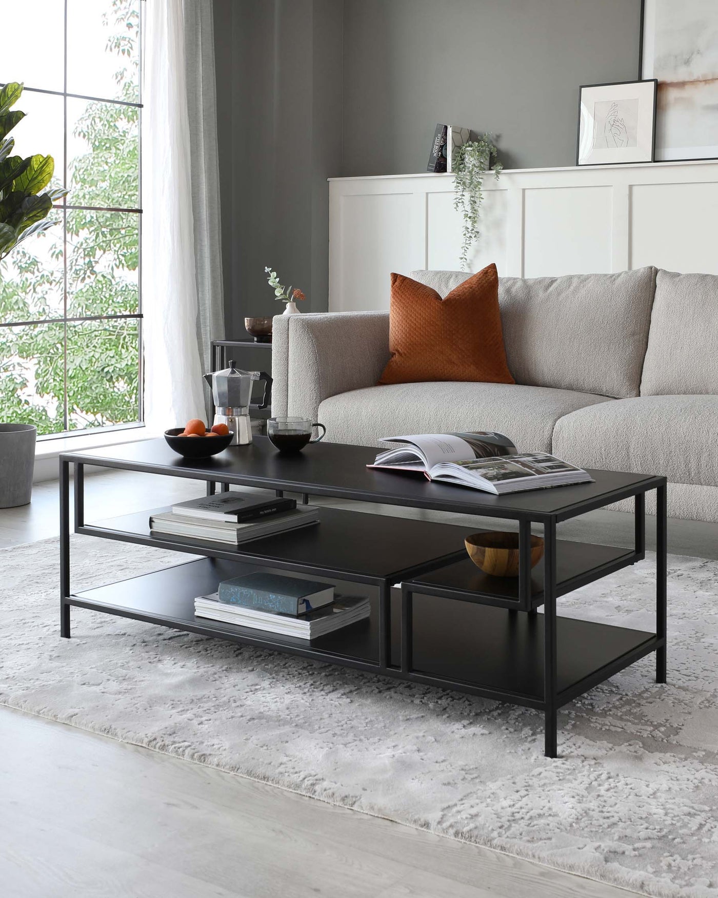 Modern living room featuring a minimalist, rectangular coffee table with a matte black metal frame and a sleek black tabletop. The table includes a lower tier with additional shelving space for books and decorative items. Gray textured sofa with a burnt orange throw pillow complements the table, both positioned on a light grey plush area rug. White credenza and artwork adorn the background, with natural light streaming through sheer curtains.