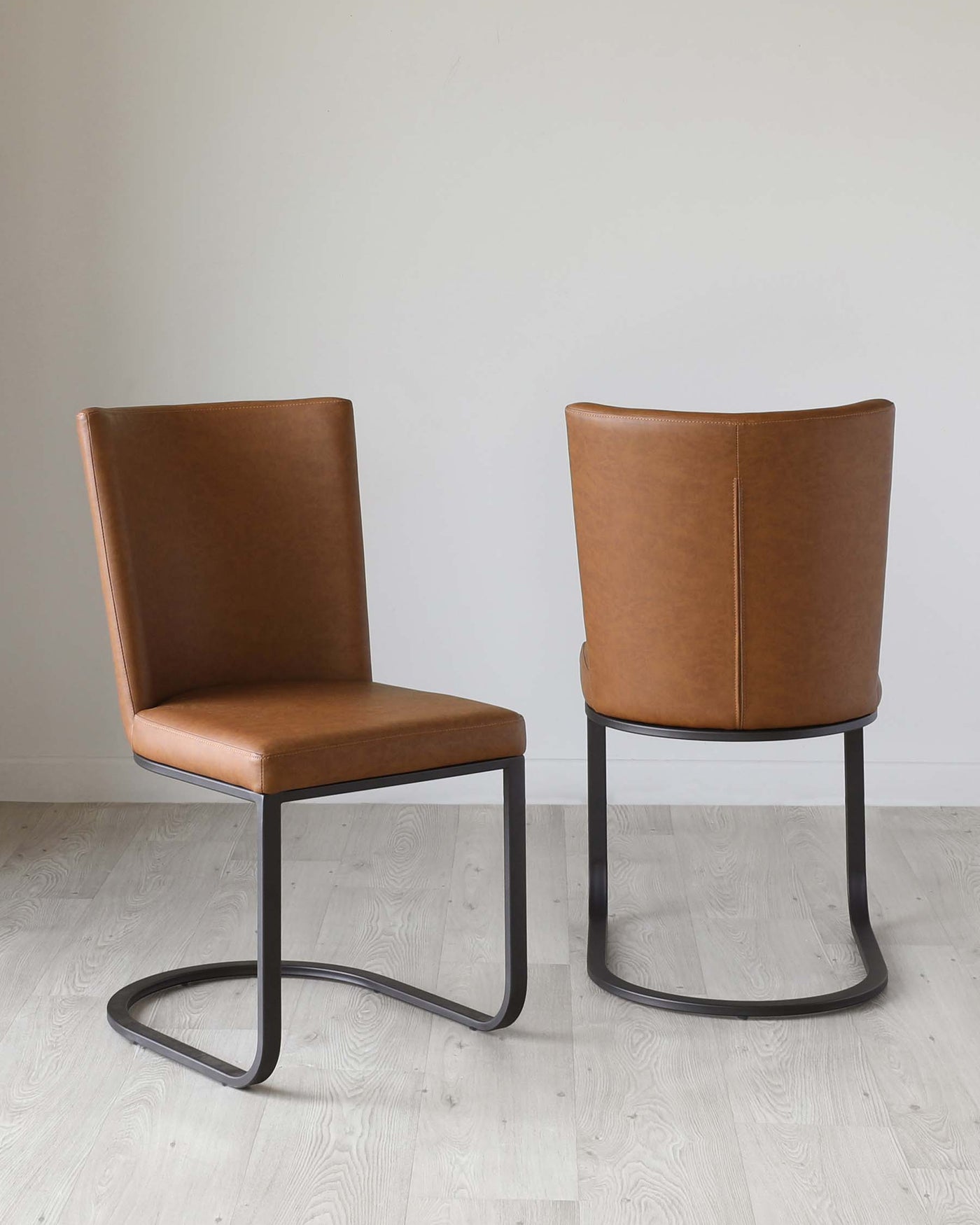 Two modern dining chairs with a sleek design, featuring caramel-coloured leather upholstery and matte black metal sled bases. The chairs have a simple yet elegant silhouette with a curved backrest for added comfort. They are placed on a light hardwood floor against a neutral wall background, highlighting their contemporary style.
