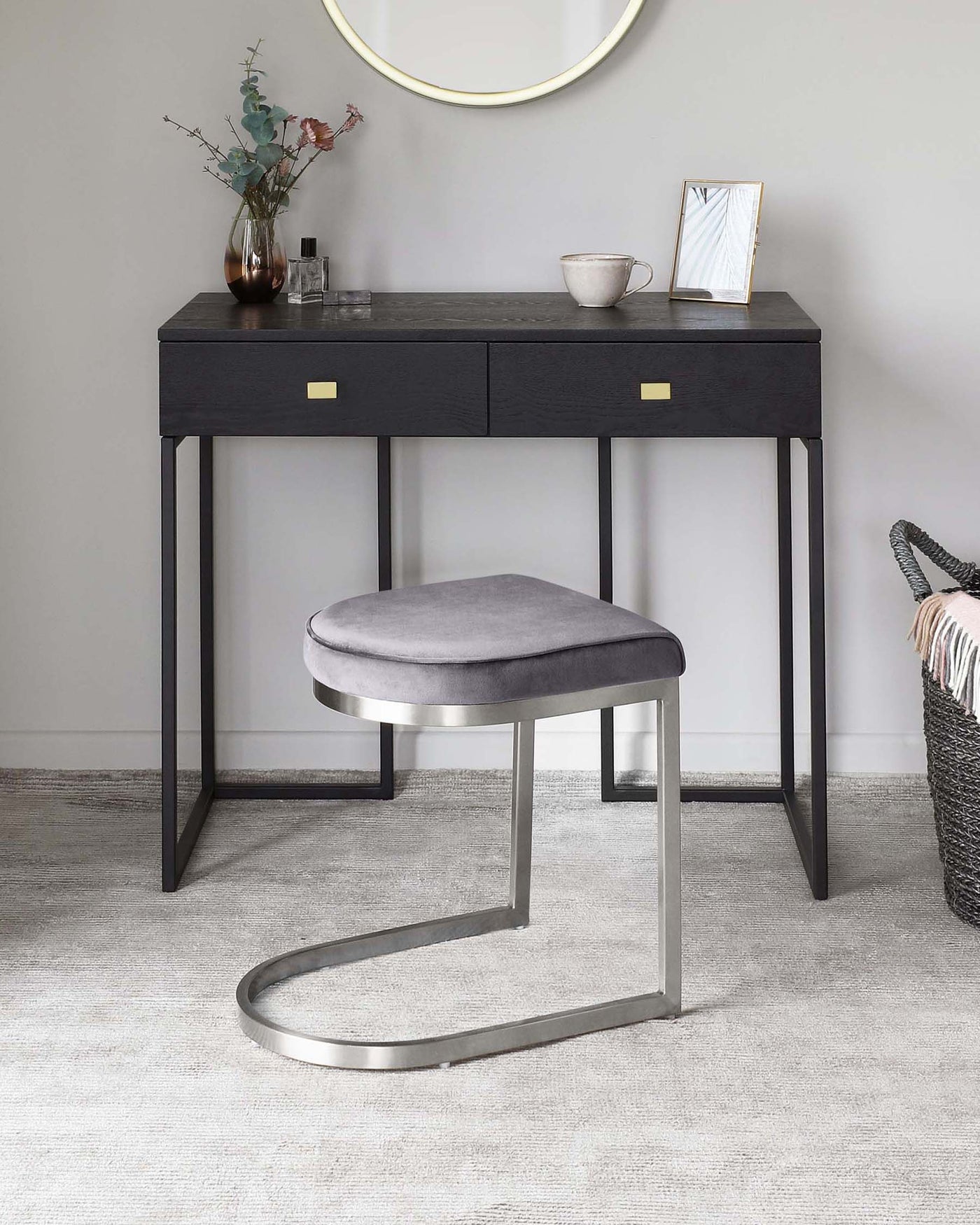A modern black wooden console table with two drawers and brass drawer pulls, paired with a round grey upholstered stool with a unique u-shaped metallic base, set in an elegant room with a light grey carpet.