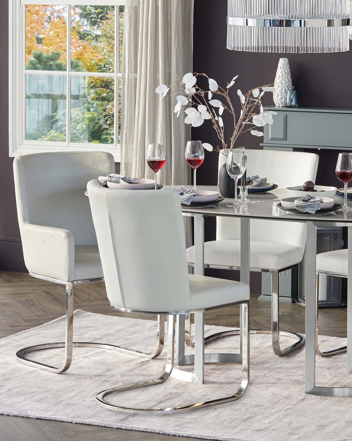 Modern dining room set featuring a rectangular table with a sleek, reflective chrome base and a clear glass tabletop. Accompanied by four elegant, high-backed chairs upholstered in white faux leather with gently curved chrome legs.