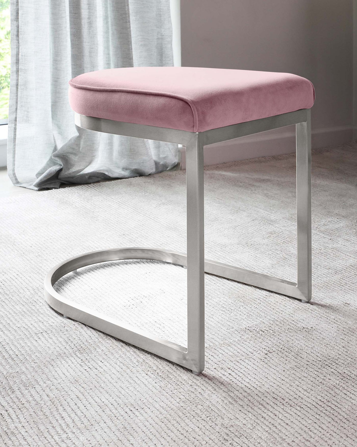 Modern minimalist stool with a plush pink upholstered top and sleek, chrome-finished metal base. The base features a simple rectangular frame with a single horizontal support bar that doubles as a footrest. The stool is set against a neutral-toned carpeted floor, complementing light-flowing curtains in the background.