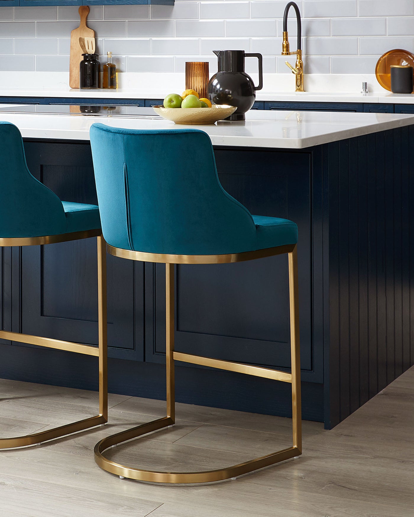 Elegant modern bar stools with plush teal upholstery and sleek gold-finished metal frames, showcased in a stylish kitchen with a navy blue island counter.