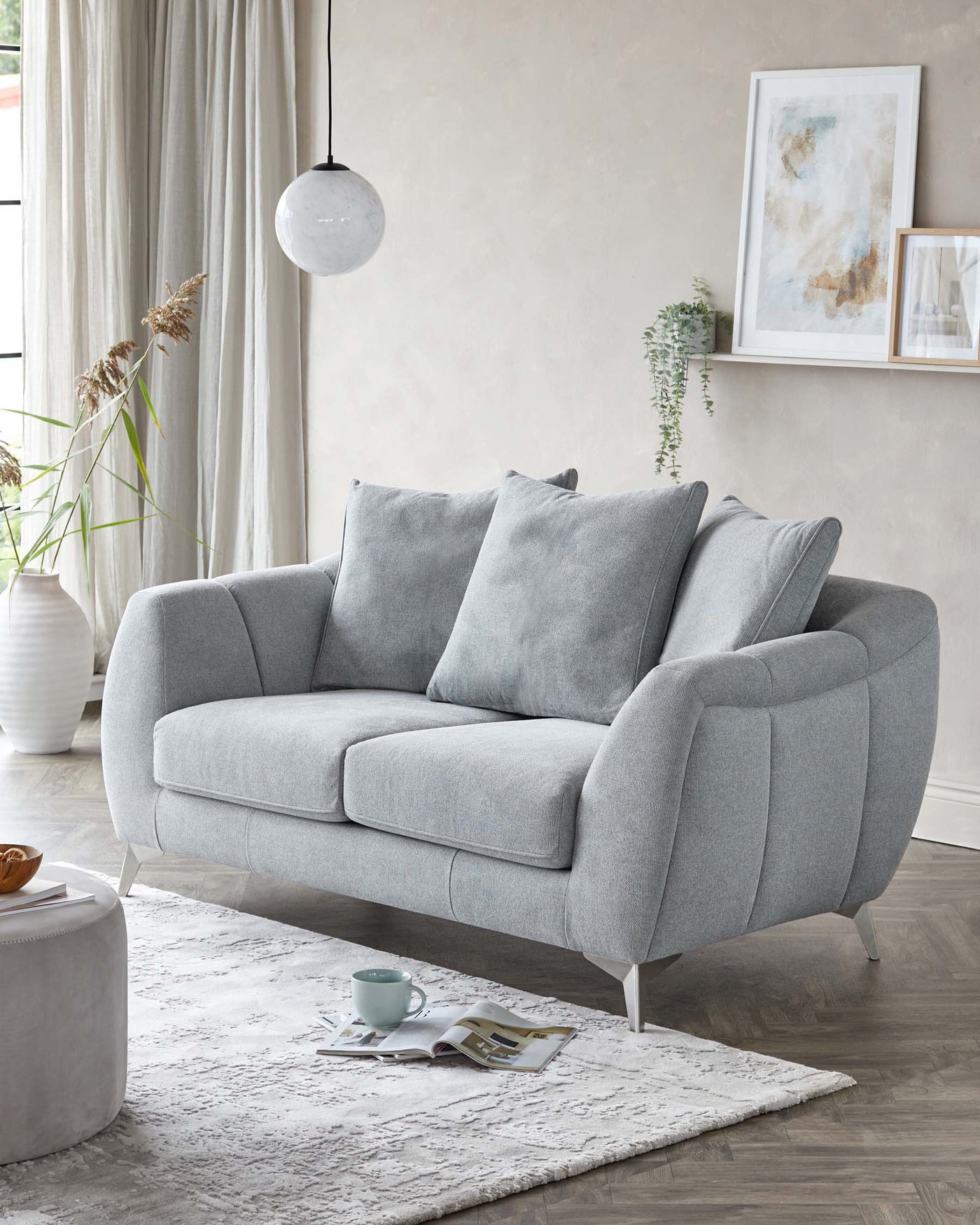 A contemporary light grey fabric sofa with a curved silhouette and sleek metal legs, paired with a rounded off-white ottoman with storage function, set upon a textured white area rug. The scene includes a minimalistic white pendant lamp and is accented with neutral-toned wall art and a green houseplant for a subtle touch of colour and life.