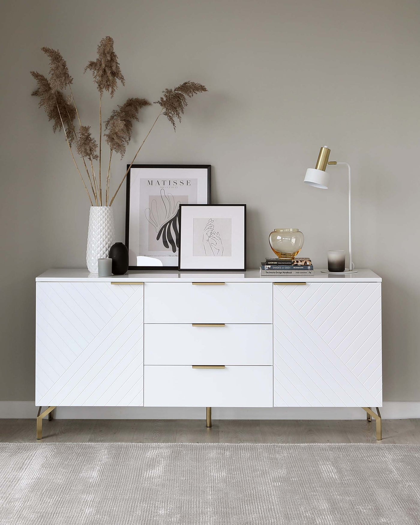 A modern white sideboard with a textured front, featuring angular lines and gold accents on the legs and drawer handles. The sideboard is adorned with decorative items including vases, framed artwork, books, and a table lamp. The scene is set against a neutral wall on top of a textured grey rug.