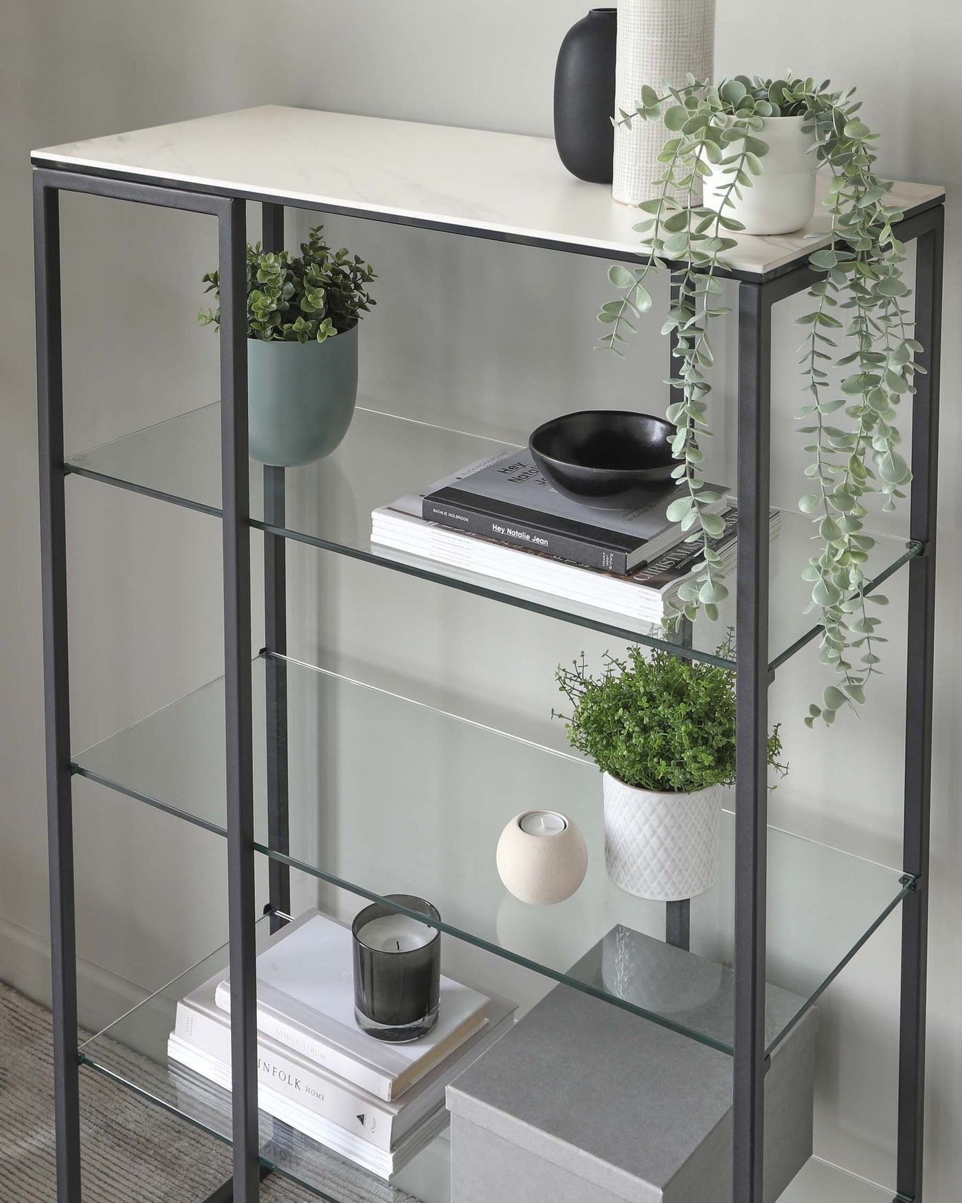 A contemporary, black metal frame bookshelf with four tiers featuring marble-like white laminate tops and clear glass shelves. Decorative items including plants, books, and vases are arranged on the shelves for display purposes.