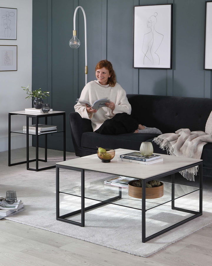 A modern living room setup featuring a sleek black metal-frame sofa with dark upholstery, a rectangular white marble top coffee table with a black metal frame, and a matching black metal-frame side table with a lower shelf displaying books and a plant. The room is decorated with subtle art and soft lighting, emphasizing the contemporary design of the furniture.