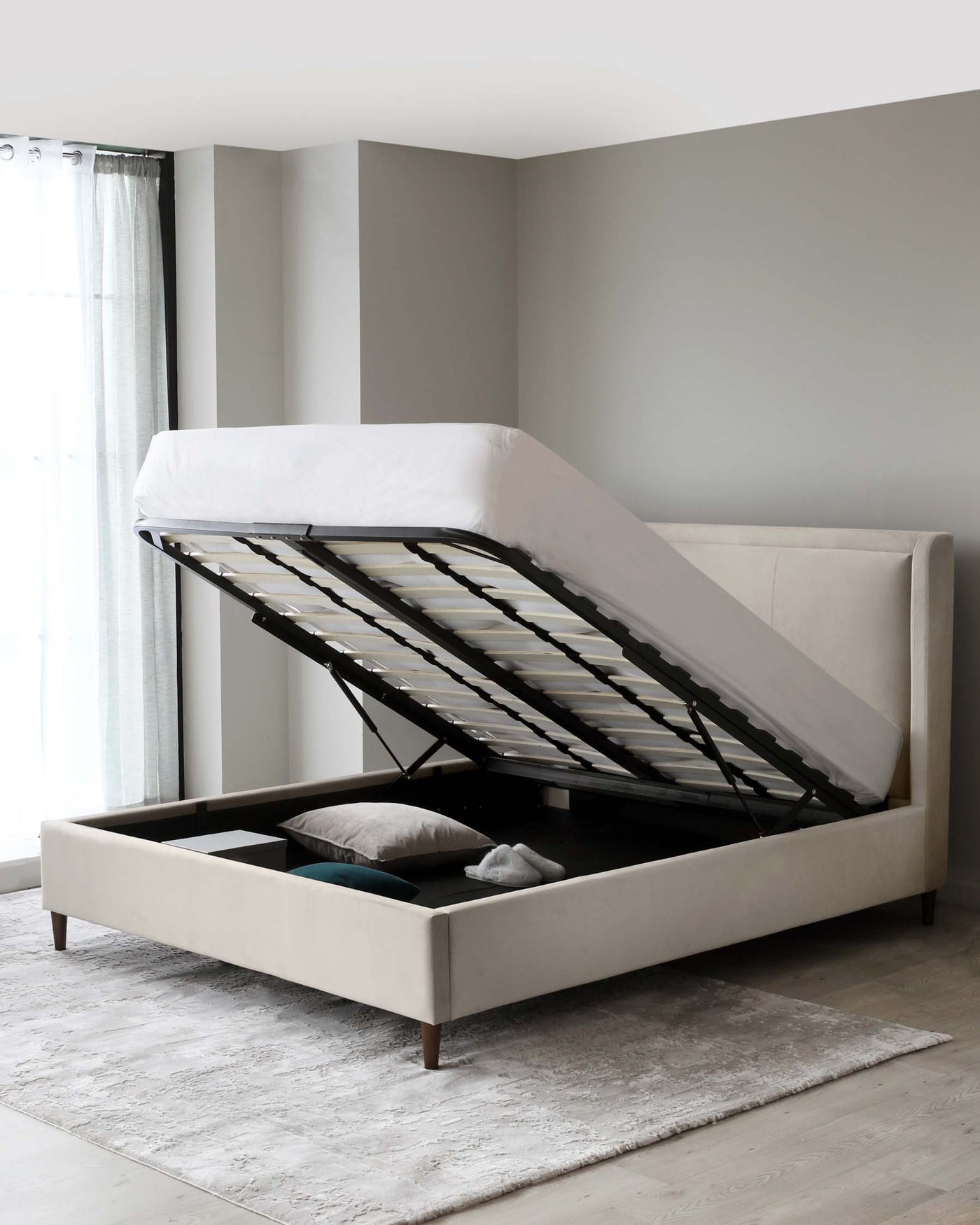 Contemporary beige upholstered storage bed with a lifted mattress revealing an organized compartmentalized space underneath. The bed features a tufted headboard and dark wooden legs, placed on a textured light grey area rug.