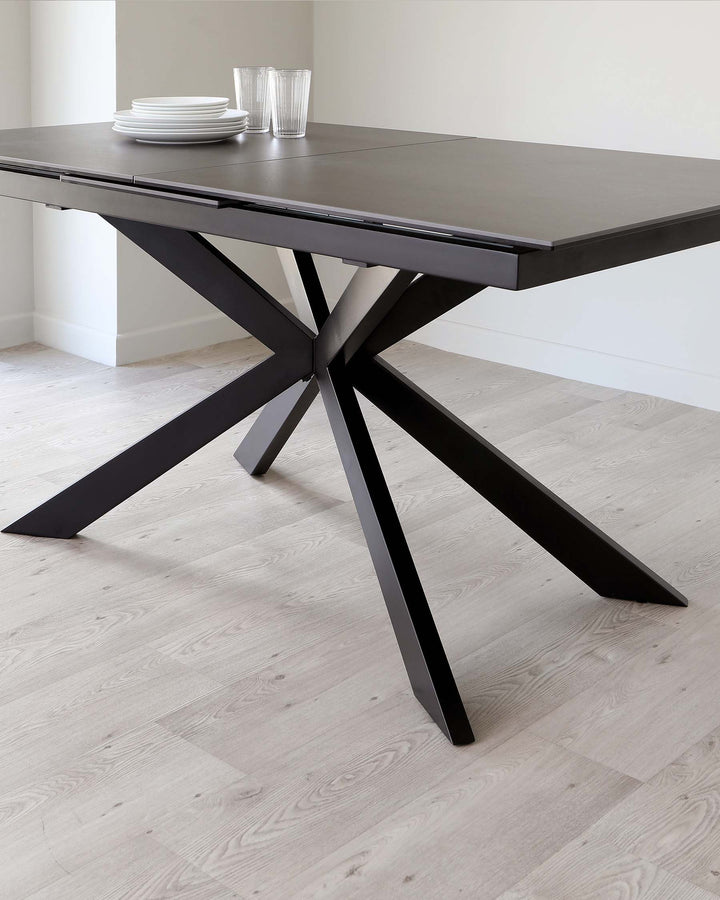 Modern dark grey extendable dining table with a unique, geometric, cross-legged base in a matte black finish, set against a light wood floor. The table is set with a small stack of white plates and clear glasses on top.