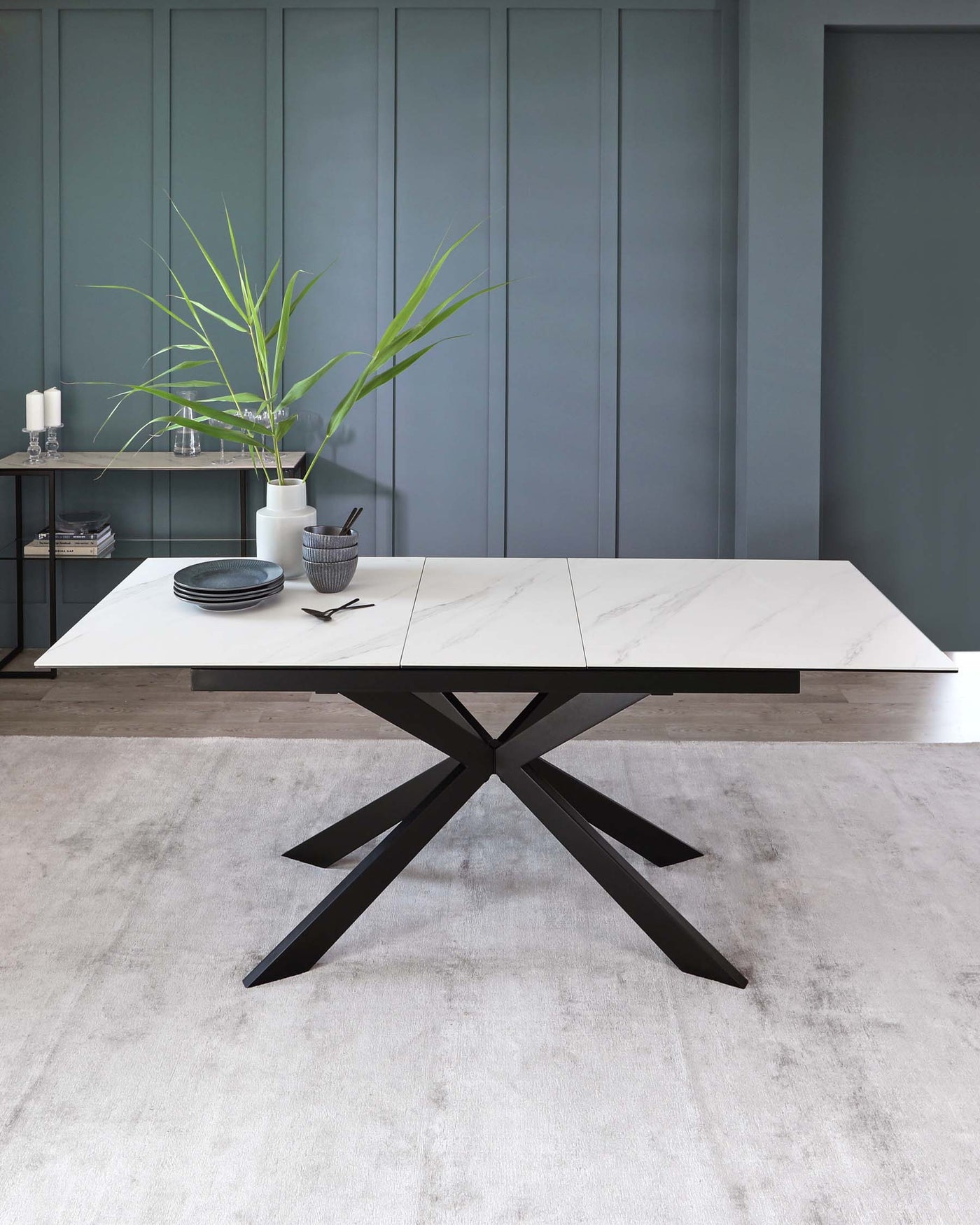 Modern rectangular dining table with a white marble tabletop and a bold black X-shaped metal base, accompanied by a minimalist metal shelf unit with decorative items against a muted teal wall.