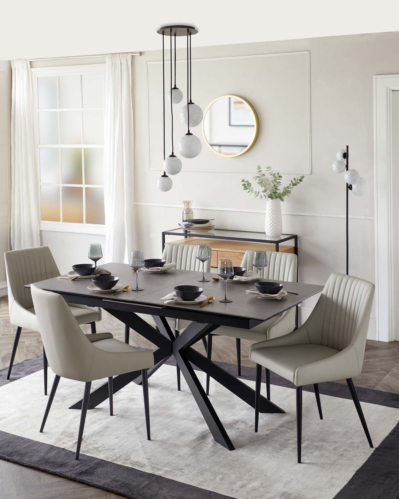 Modern dining room set featuring a rectangular black table with a unique, angular black base design. The table is surrounded by six beige upholstered chairs with vertical channel tufting on the backrests and slim black legs. A simple black sideboard is situated against the wall, accented with decorative items. The furniture is arranged on a textured grey area rug.
