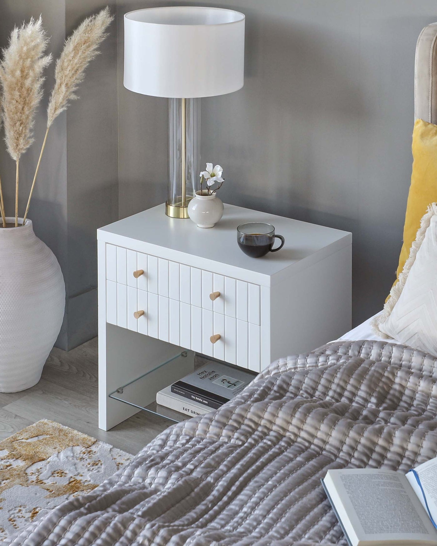 Modern white bedside table with three drawers featuring wooden knob handles and an open lower shelf displaying books, paired with a large white lamp with a transparent stand and brass accents.