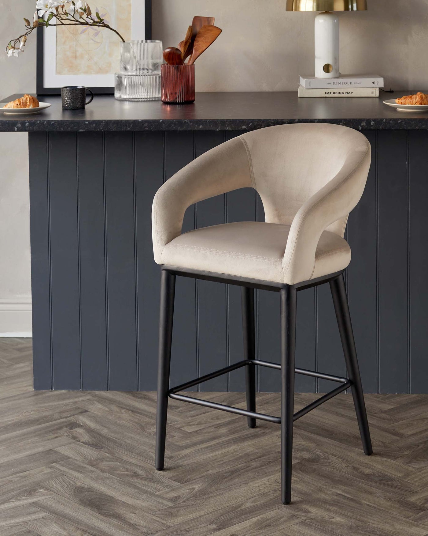 Elegant modern bar stool with a curved backrest and plush beige upholstery, featuring a sleek black metal frame with four legs and a footrest.