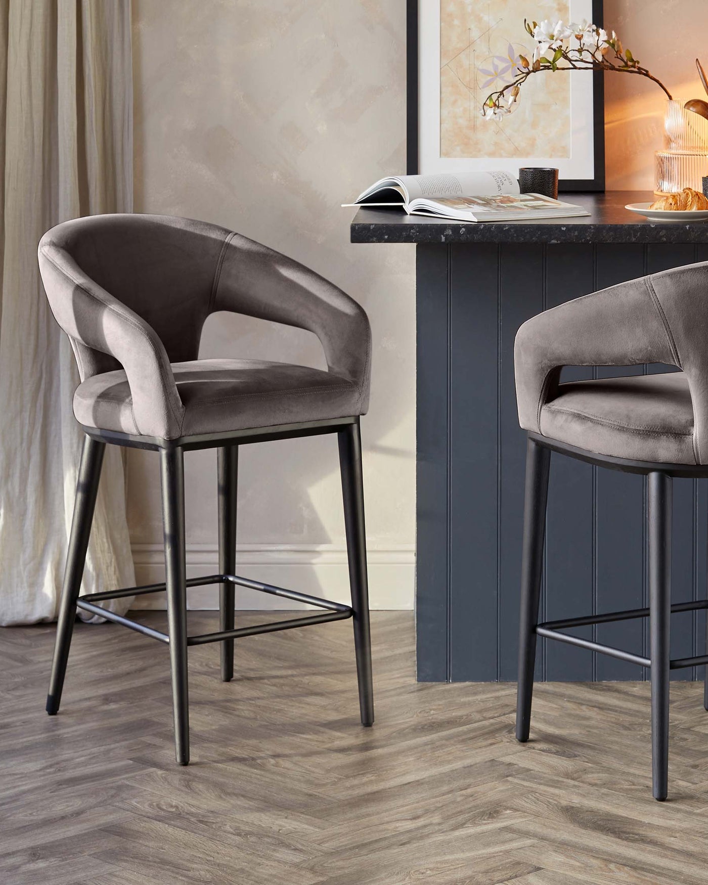 Elegant pair of grey velvet bar chairs with a smooth curved backrest, comfortable armrests, and slender black metal legs with footrests, showcased in a stylish interior setting.