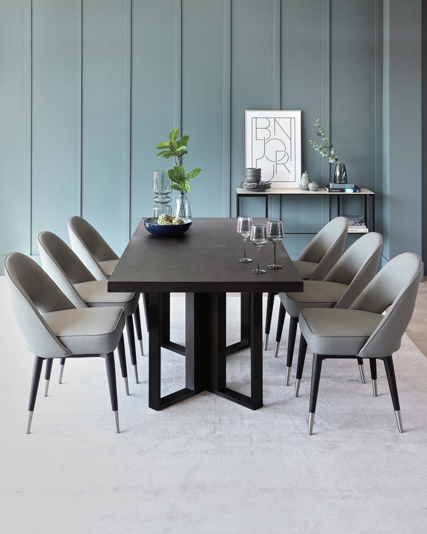 Modern dining room featuring a large rectangular dark wood table with robust, block-style legs, surrounded by eight elegant light grey upholstered dining chairs with slender metal legs. A sideboard with decor accents is placed against a panelled wall in the background.