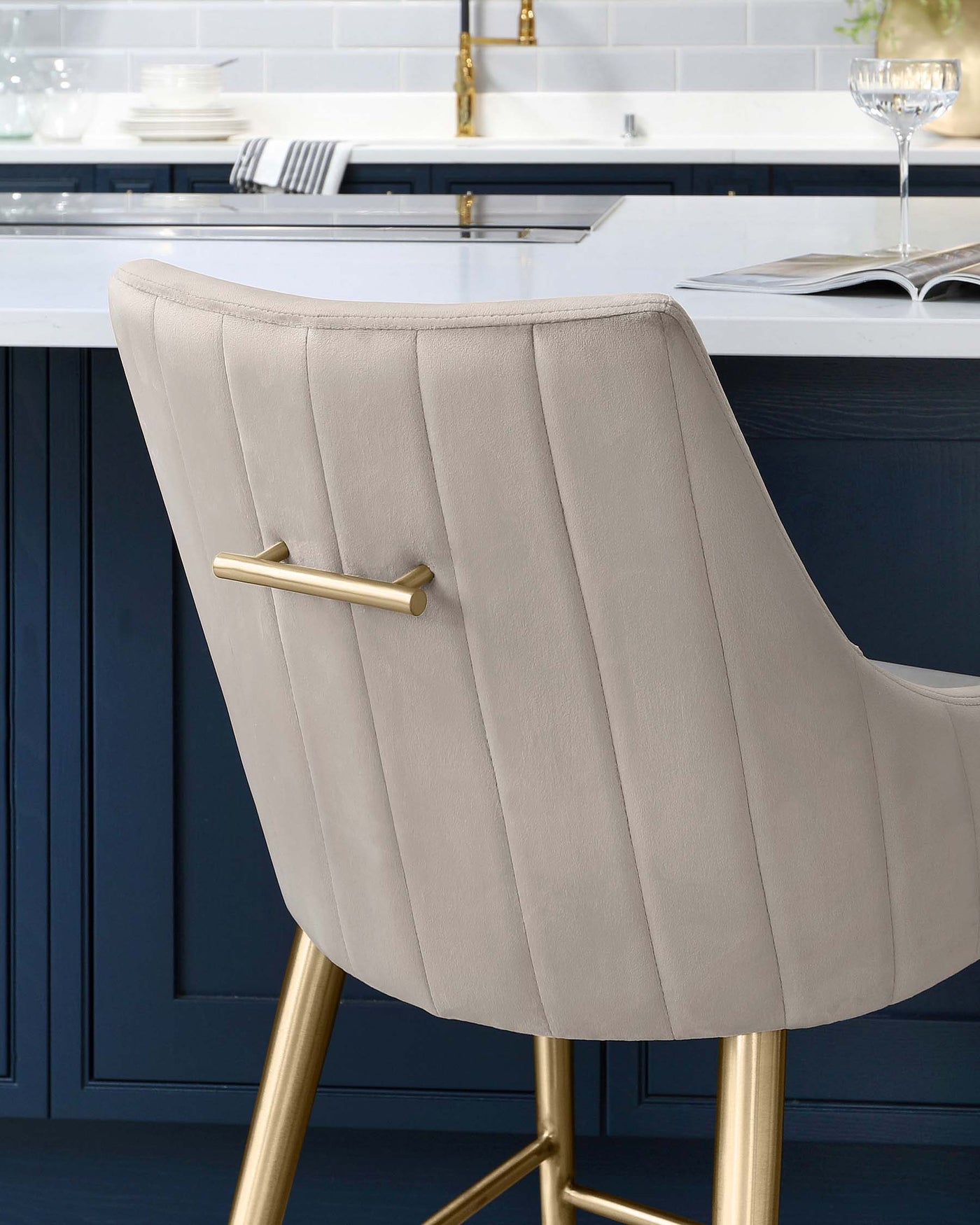 Elegant modern bar stool with beige upholstery and vertical channel stitching. The stool features a curved backrest and a gold-finished metal frame with four sleek legs that intersect at the base, providing sturdy support.
