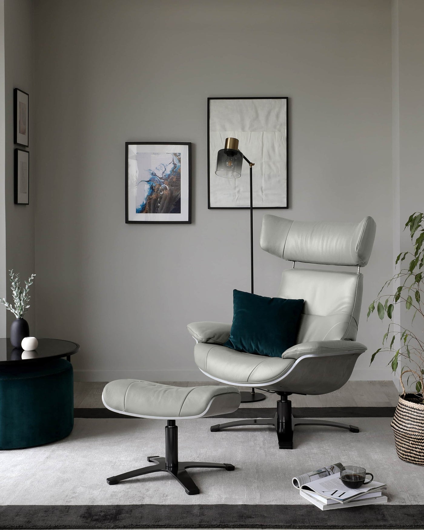 Elegant modern furniture set in a minimalist living room, including a light grey leather recliner chair with a matching footstool, both featuring sleek black metal bases. A round, dark blue velvet ottoman sits beside a matte black coffee table with a white vase and greenery. The room is decorated with framed artwork, a floor lamp, and accent cushions.
