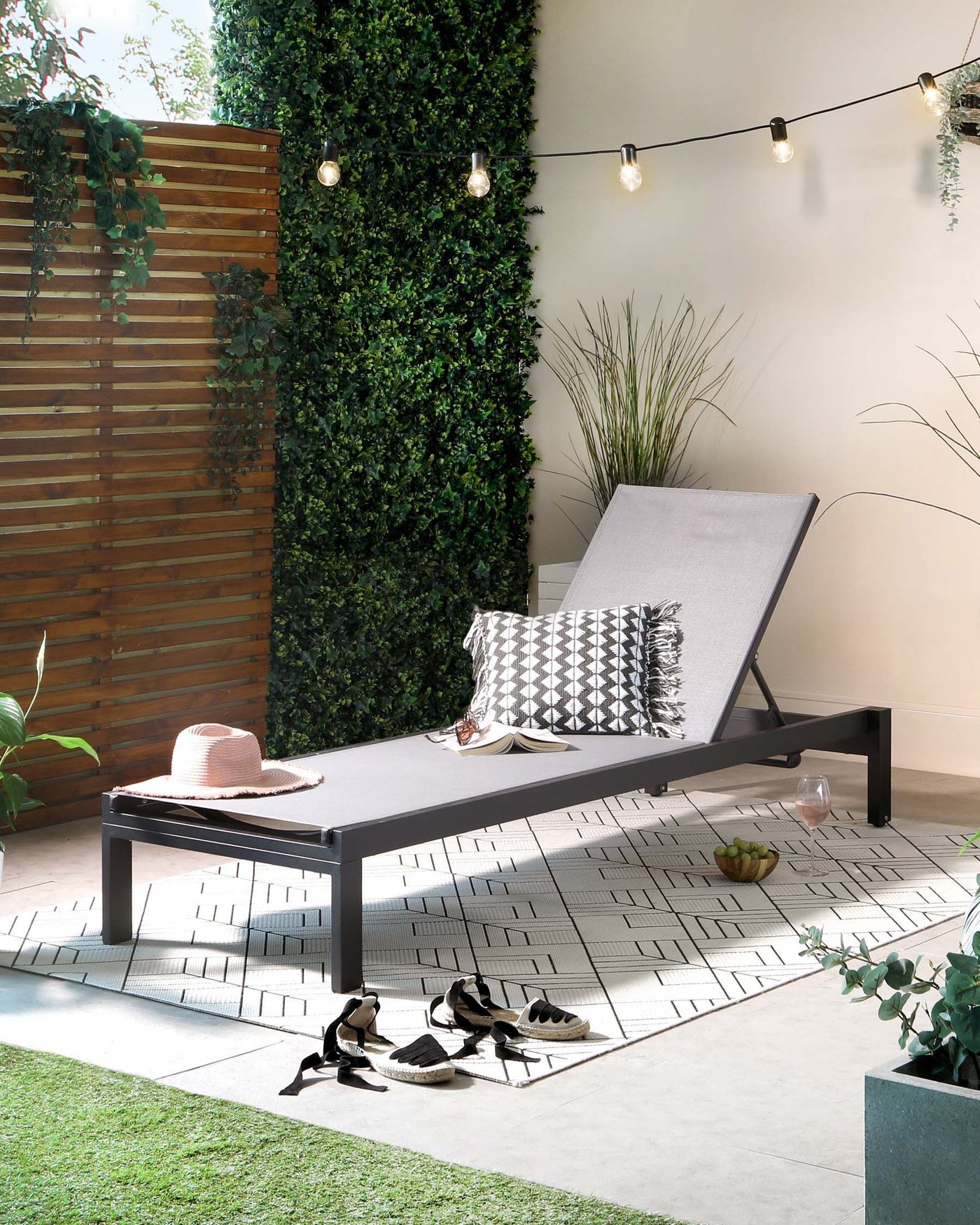 Modern outdoor chaise lounge in dark grey with an adjustable backrest and a textured grey cushion, accented with a black and white geometric patterned throw pillow and a fringed blanket.