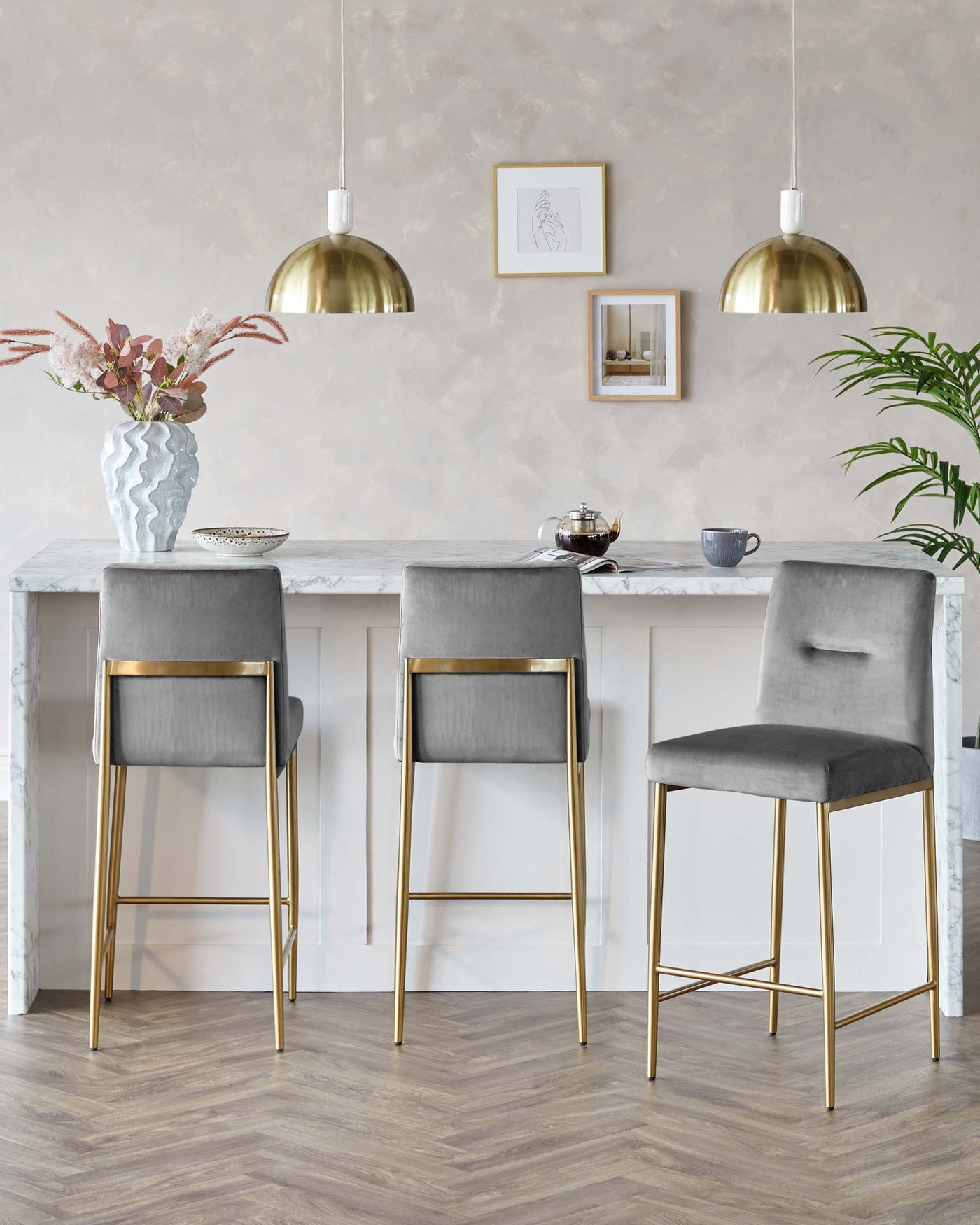 Modern elegant kitchen set with three grey upholstered bar stools featuring gold metal legs, placed at a white kitchen island with a marble countertop.