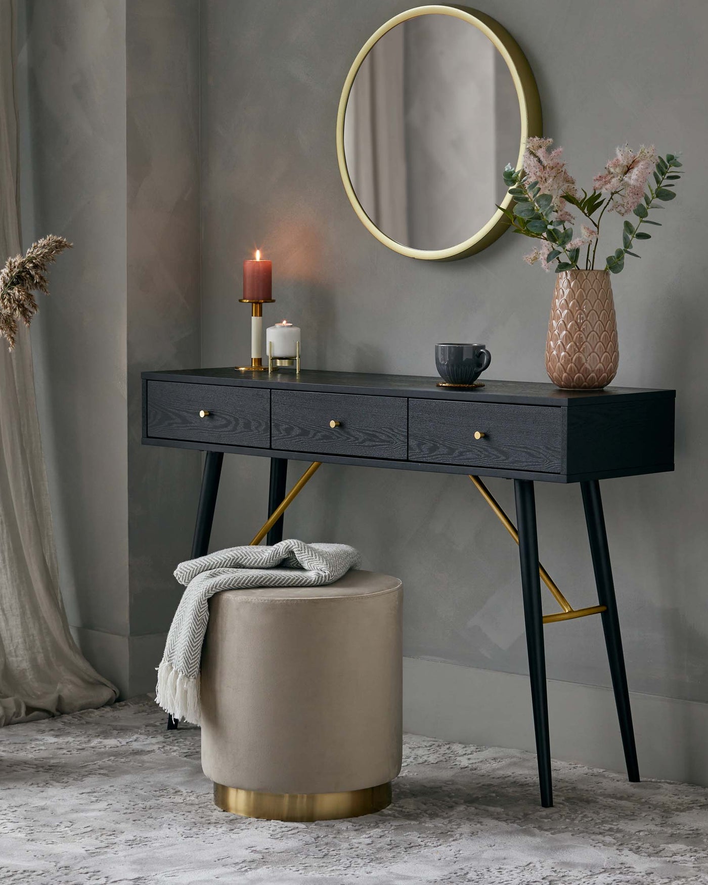 Elegant modern furniture set against a grey textured wall, featuring a sleek black wooden console table with brass handles and slim tapered legs with brass accents, paired with a round, beige upholstered ottoman with a brass base. A tasteful arrangement of accessories complements the scene, including a round mirror with a brass frame, decorative vase with flowers, candles, and a cosy throw.
