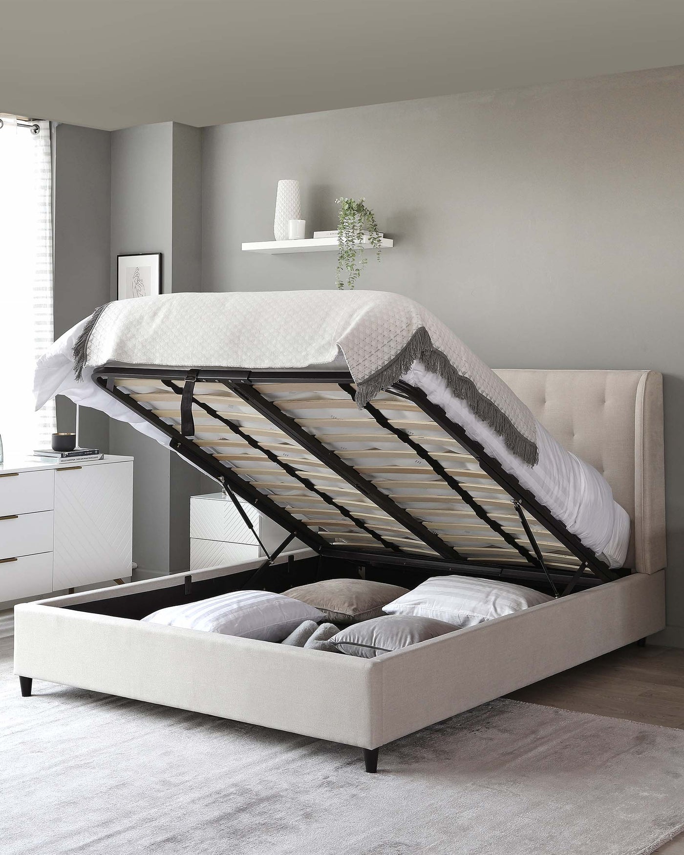 Beige upholstered storage bed with a lifted mattress platform revealing ample storage space underneath, paired with a modern white bedside drawer unit with a black lamp and matching dresser in a grey bedroom setting.