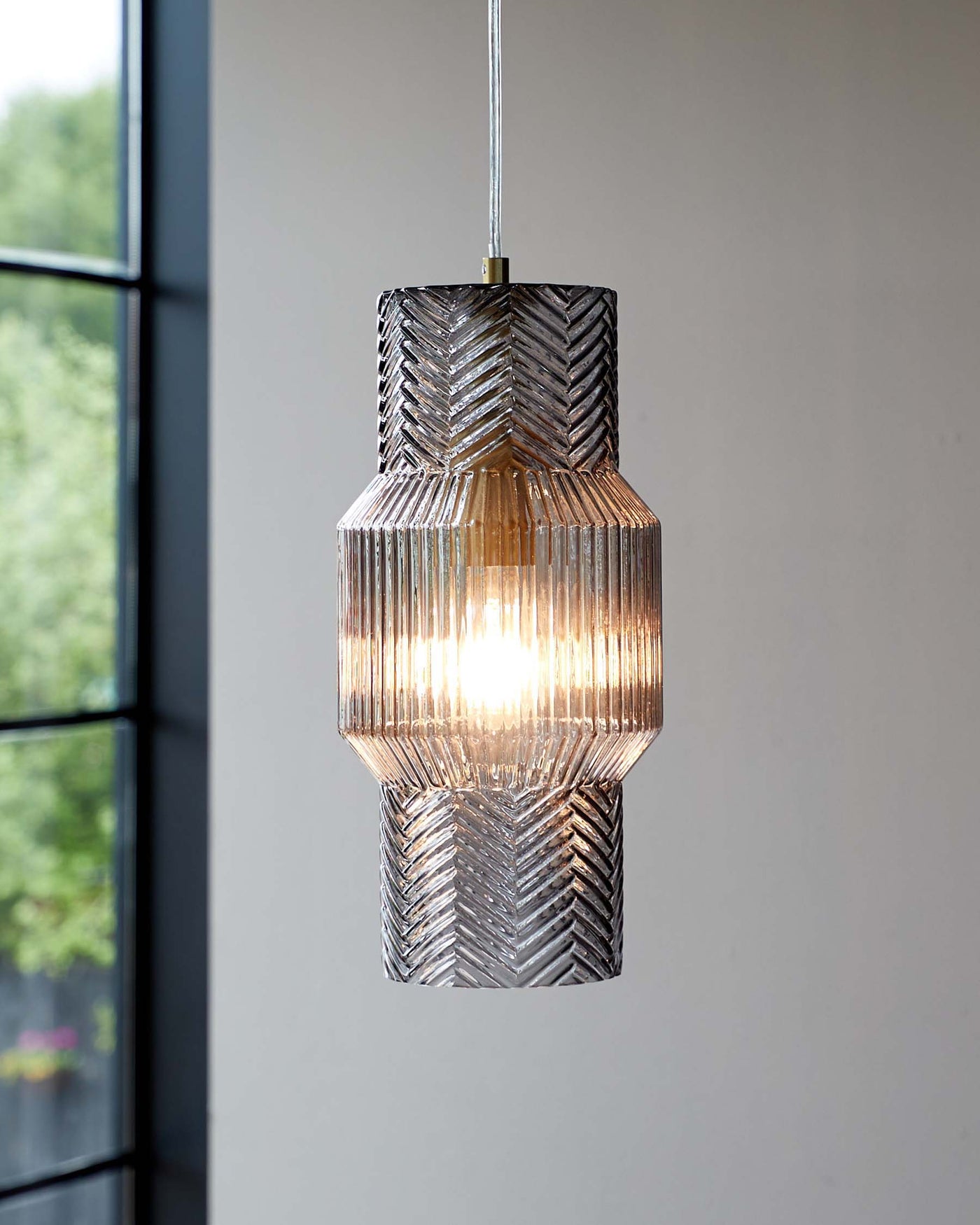 Elegant pendant light with a textured glass design comprising multiple angular ridges, creating a sparkling lighting effect. The fixture features two distinct sections, a smaller top part atop a larger bottom section, both with a matching geometric pattern. A warm glow emanates from within, highlighted by the glass's ridges. The light suspends from a metallic cord, complementing the modern and sophisticated look, perfect for a contemporary interior setting.