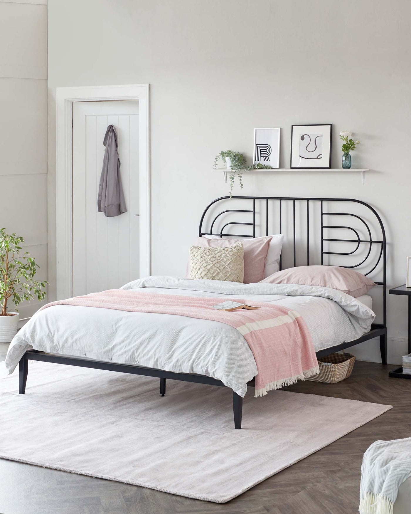 Modern minimalist bedroom with a black metal bed frame featuring a curved headboard, set against a light grey wall. The bed is dressed with white and blush pink bedding, accented by decorative pillows. A light pink area rug rests beneath the bed, and a white floating shelf above the headboard displays framed artwork. A woven basket sits at the bed's foot on the rug, softening the overall contemporary aesthetic.