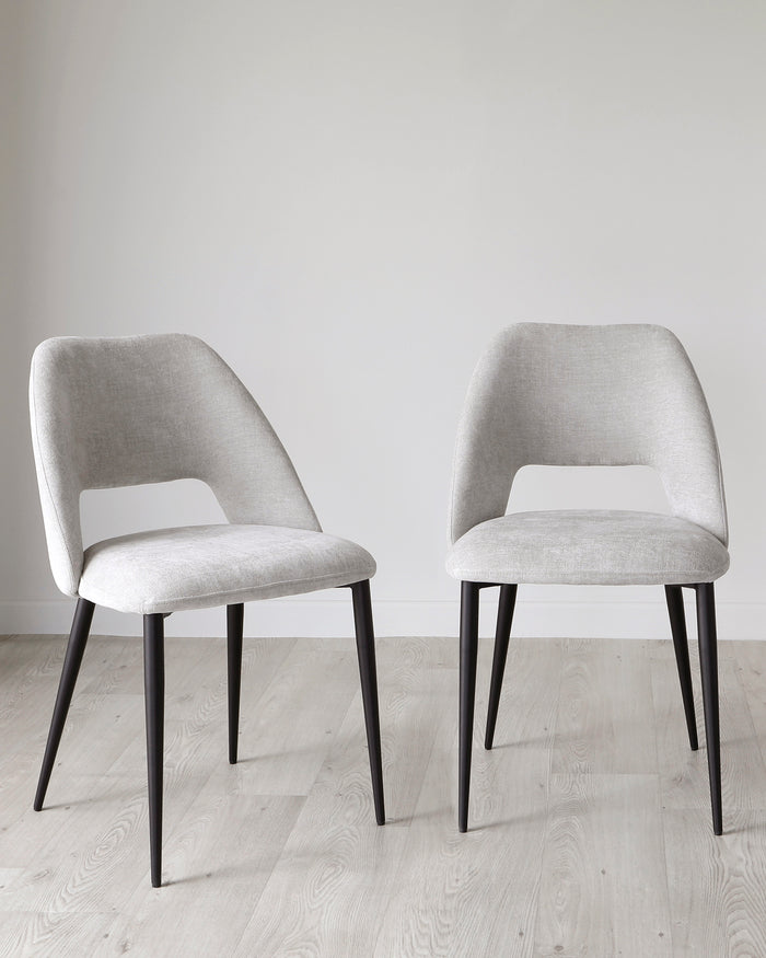 Two modern upholstered dining chairs with curved backrests and black tapered legs on a light wooden floor, against a neutral background.
