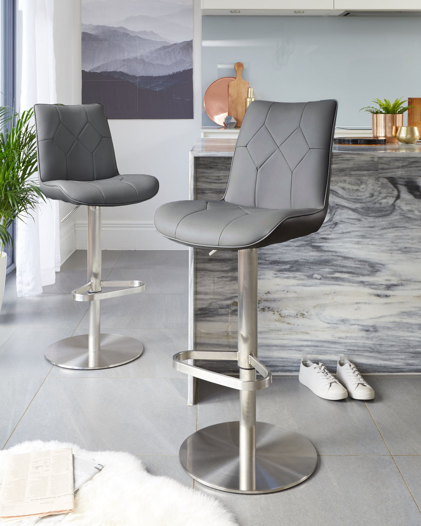 A pair of modern bar stools with chrome pedestals, featuring adjustable height, a footrest, and quilted faux leather seats in a neutral grey tone, suitable for contemporary kitchen or bar areas.
