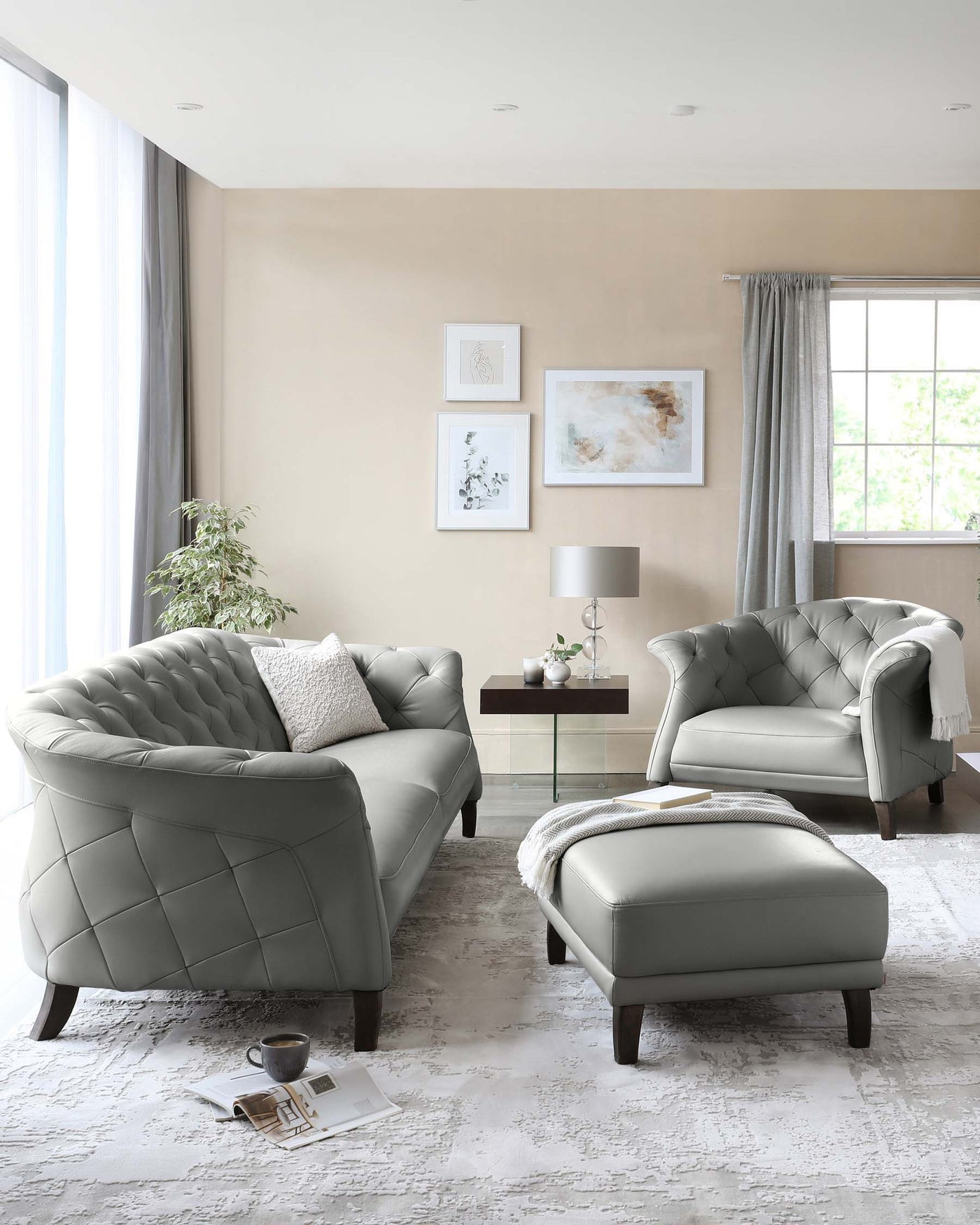 A contemporary living room set featuring a plush, tufted, light grey leather sofa and a matching armchair with quilted detailing. Between them stands a small square, dark-stained wooden coffee table with a glass vase and a pair of modern table lamps. To the right, a coordinating ottoman complements the set, draped with a soft white throw blanket.