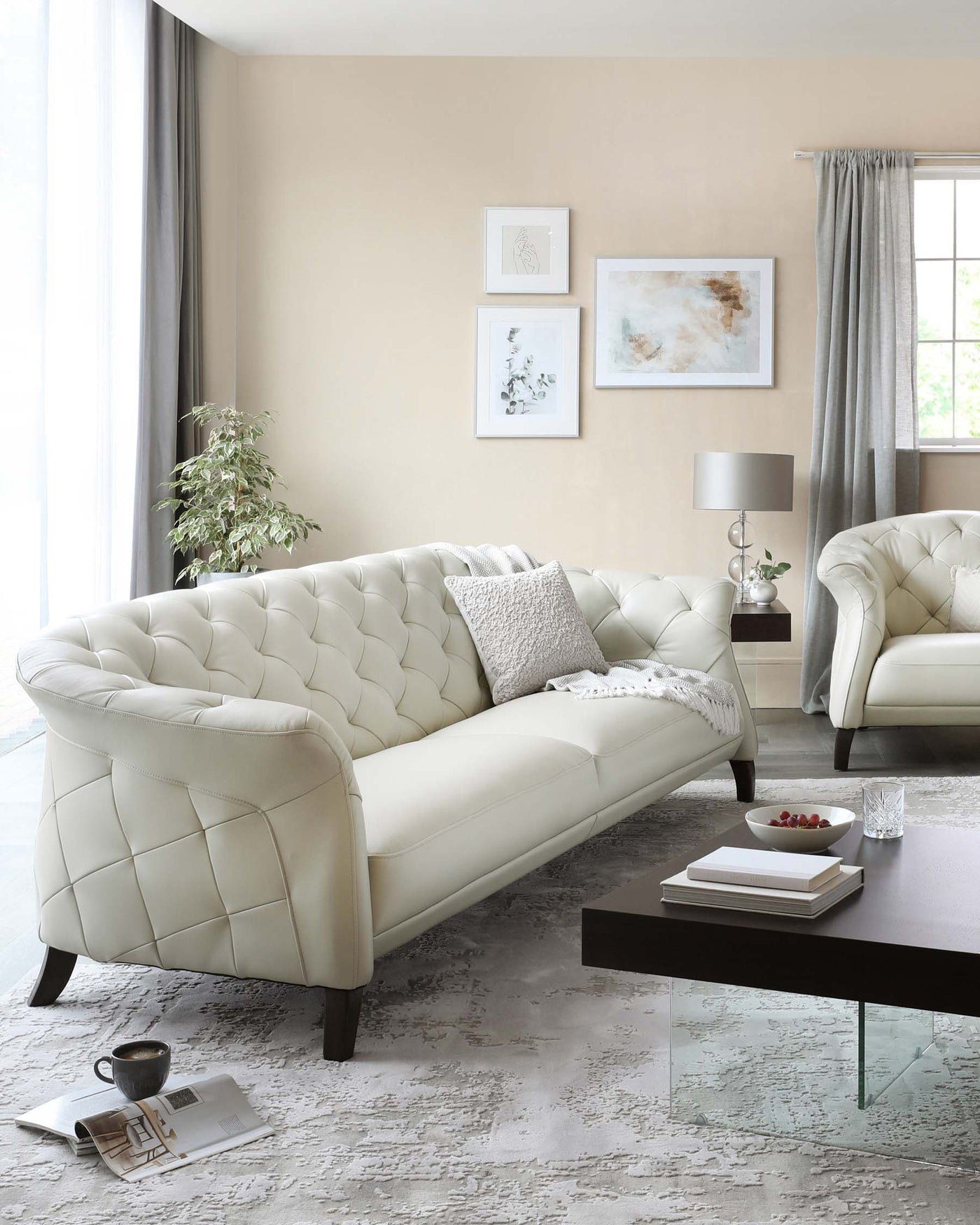 Elegant living room set featuring a tufted cream-colored leather sofa and matching armchair, complemented by a rectangular dark wood coffee table with a clear glass top. A textured off-white area rug underlies the furniture arrangement.
