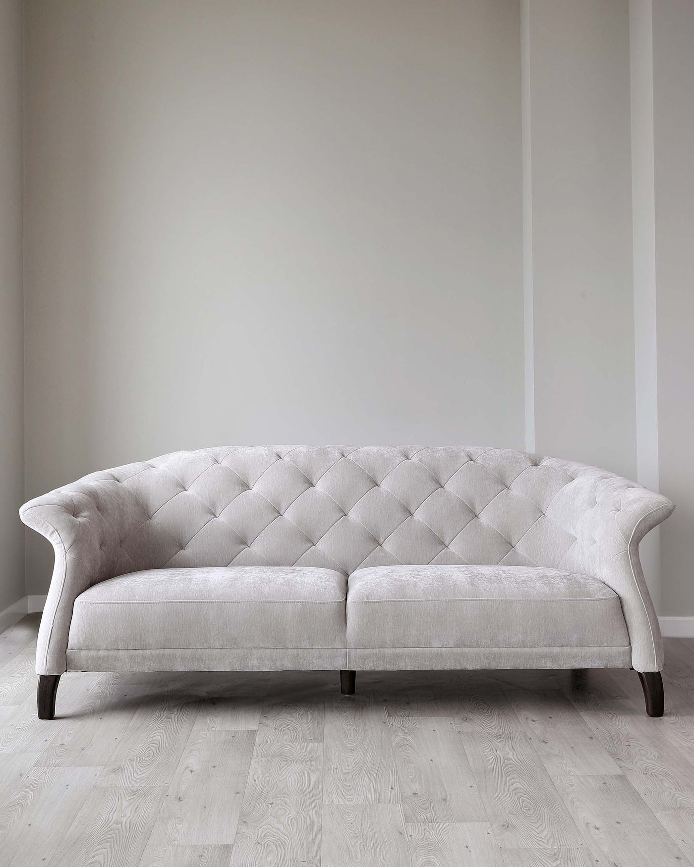Elegant light beige Chesterfield sofa with tufted upholstery and rolled arms, featuring dark wooden legs, positioned on a light hardwood floor against a neutral wall.
