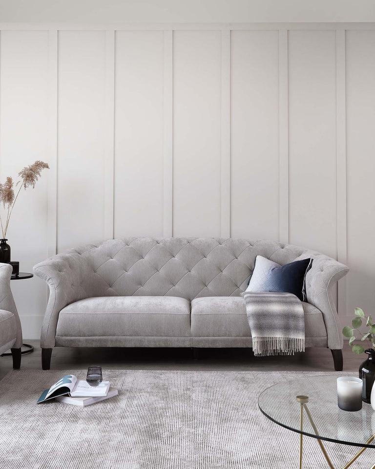 Elegant light grey tufted fabric sofa with rolled arms and a curved, button-tufted backrest. Two round side tables in black, one with a vase of pampas grass, the other with books and a glass. A grey woven area rug underfoot and a round coffee table with a gold frame and glass top complete the scene.