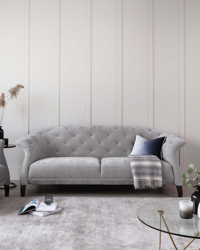 Elegant grey tufted chesterfield sofa with plush upholstery, accompanied by a round gold and glass coffee table, resting on a textured off-white area rug. Decorative accents include a dark side table with vase, books, candle, and a cosy throw blanket draped over the sofa.