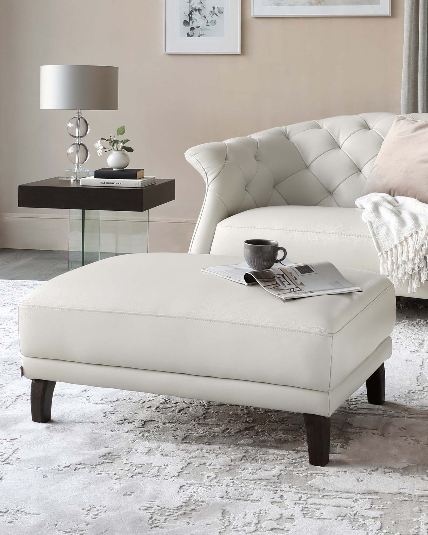 Elegant living room setup featuring a tufted cream-colored sofa with a high back and rolled arms, a matching rectangular ottoman with a smooth surface and wooden legs, and a modern side table with a black glossy finish and glass top, accentuated by a contemporary silver table lamp, decorative vase, and assorted books.