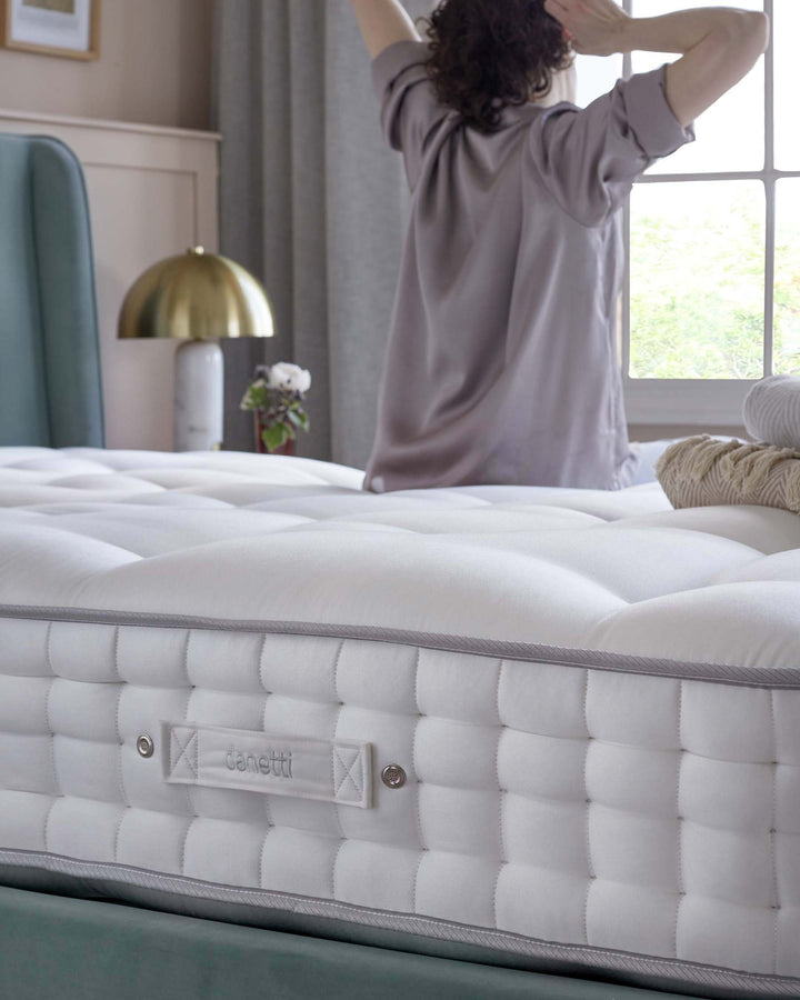 A luxurious tufted mattress with a plush, pillowy surface. The side of the mattress features stitched padding design with a branded tag and two buttons adding a detailed touch. The mattress rests on a teal bed frame that's partially visible. In the background, a gold table lamp and a vase with flowers on a white bedside table complement the elegant bedroom decor.